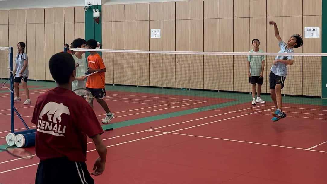 Several kids playing badminton on an indoor red court, one player jumping to smash the shuttlecock over the net.
