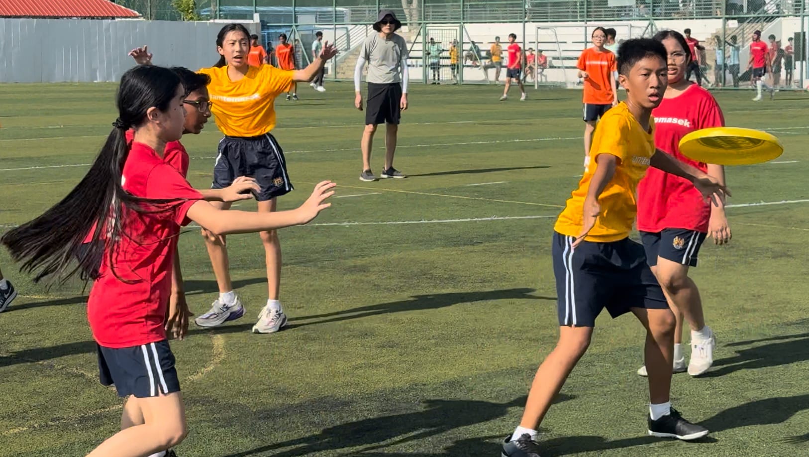 Students in red and yellow jerseys play ultimate frisbee on a green field, with one boy holding a yellow disc.