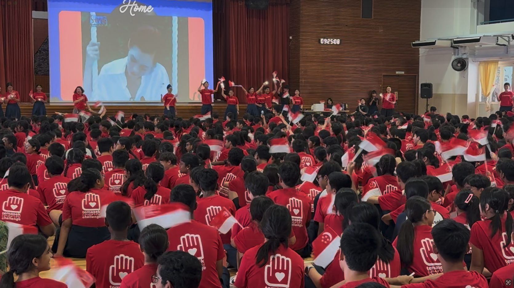 Large group of students in red Temasek charity shirts waving Singapore flags in a school hall with a projection screen.