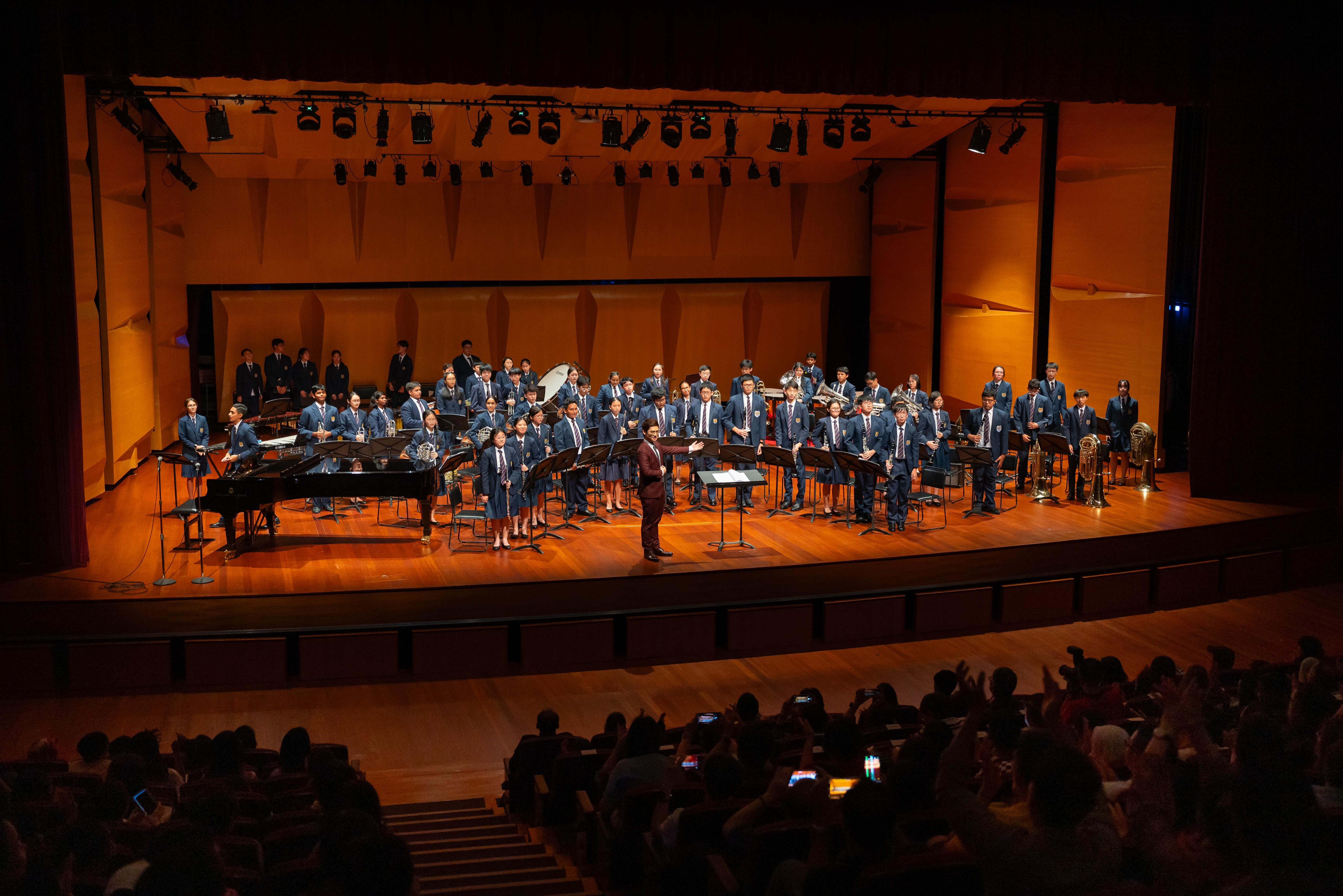 A large student band in navy uniforms stands on a warmly lit concert hall stage with a conductor and audience.