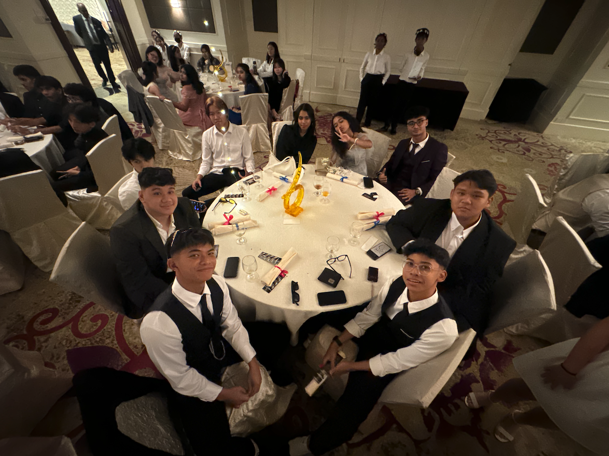 Young people in formal attire sit around a round banquet table in a decorated ballroom with more guests in background.