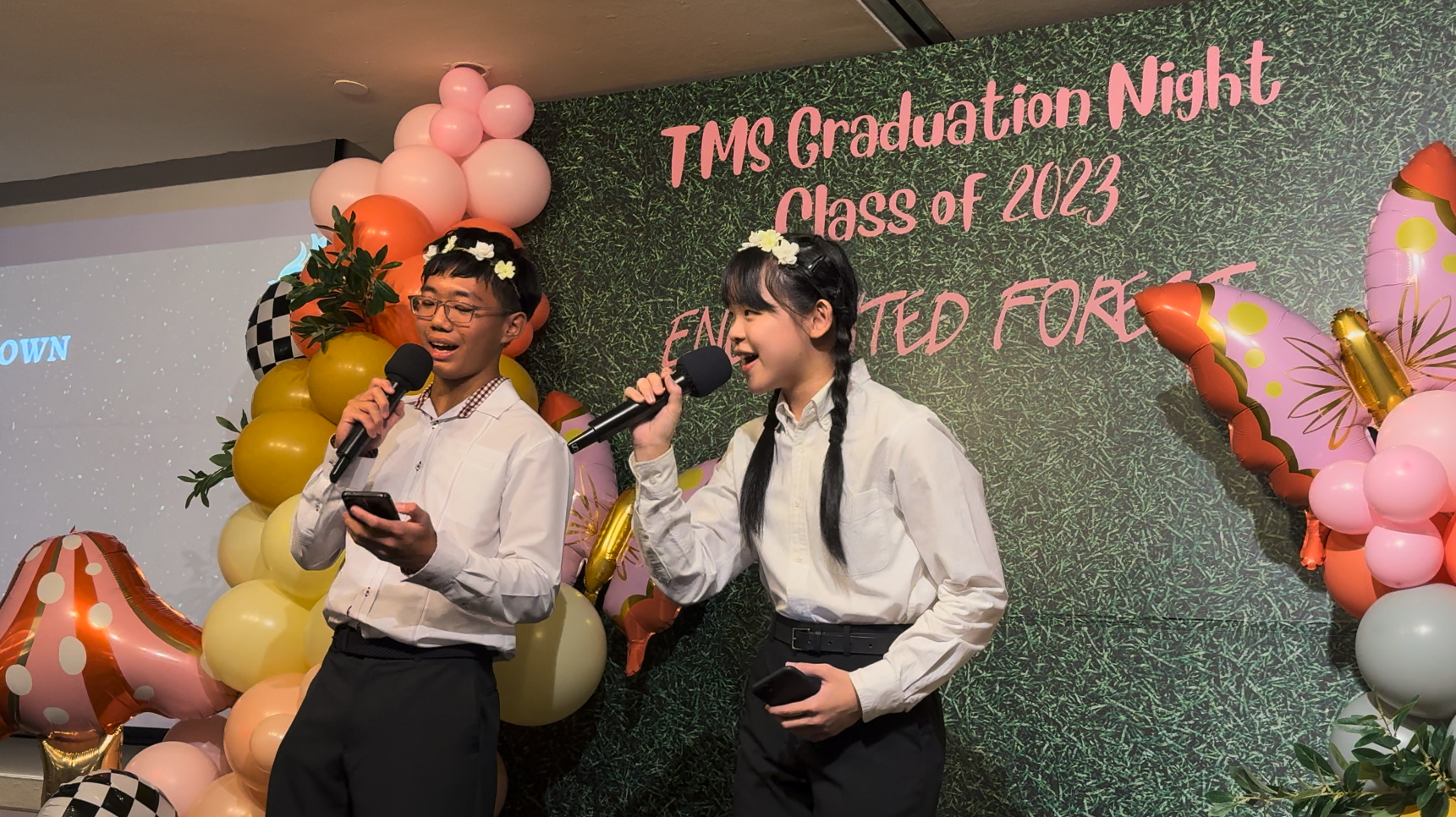 Two students speak into microphones at TMS Graduation Night Class of 2023 event, surrounded by colorful balloons.