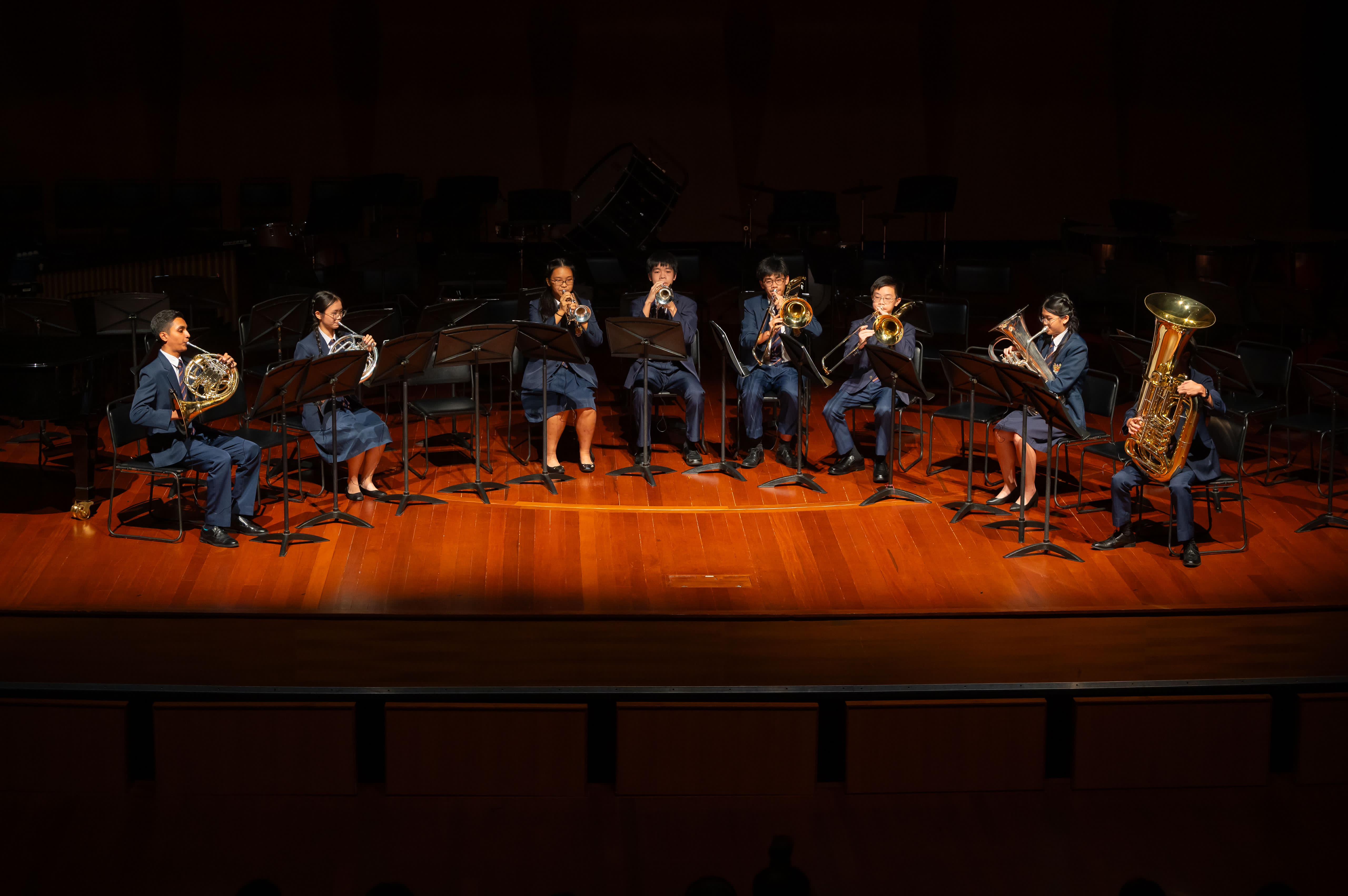 Seven young musicians in blue uniforms play brass instruments on a lit wooden concert stage with music stands.