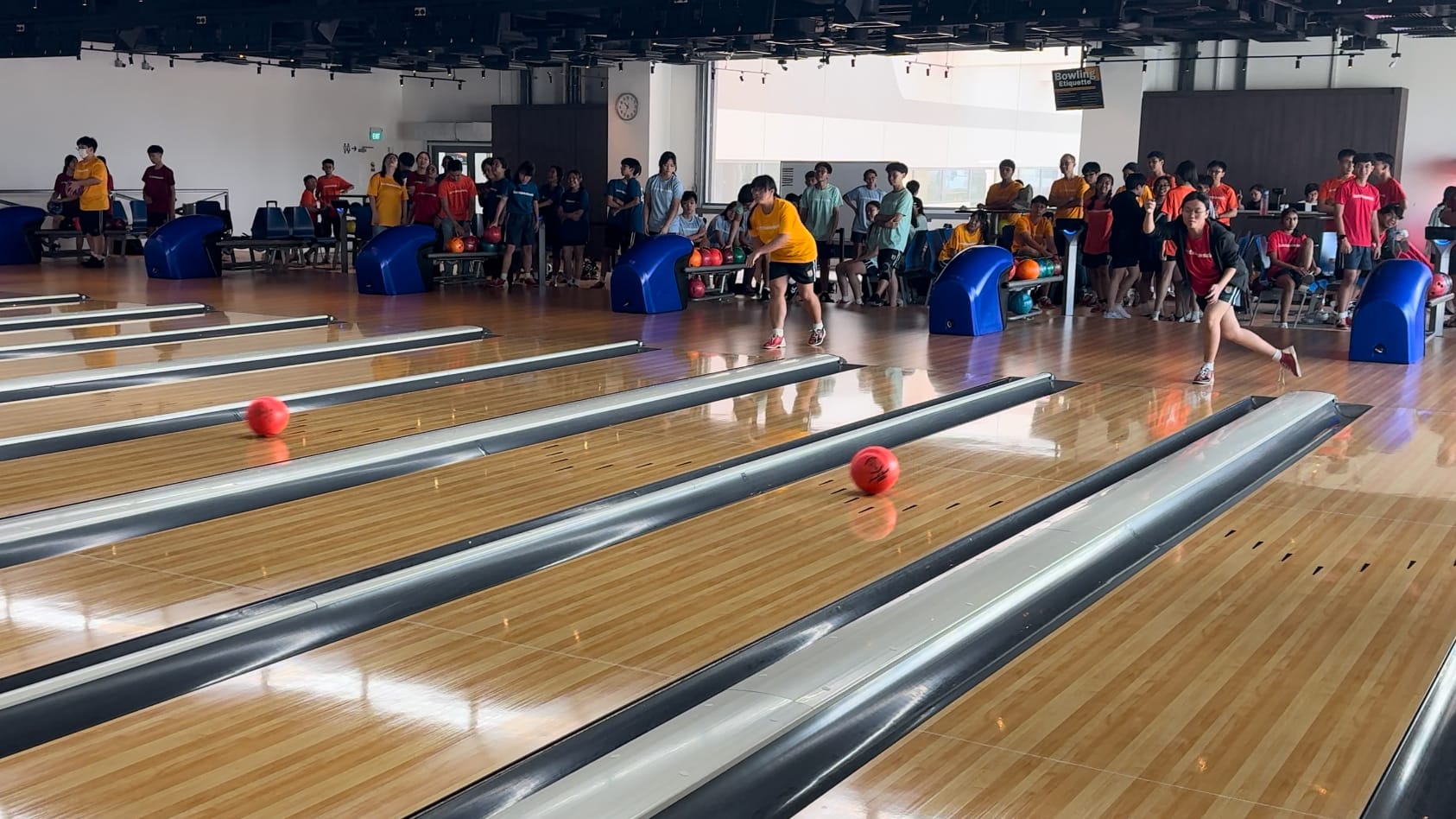 Two bowlers release red balls on polished lanes while a large crowd of young people watches from behind the ball returns.
