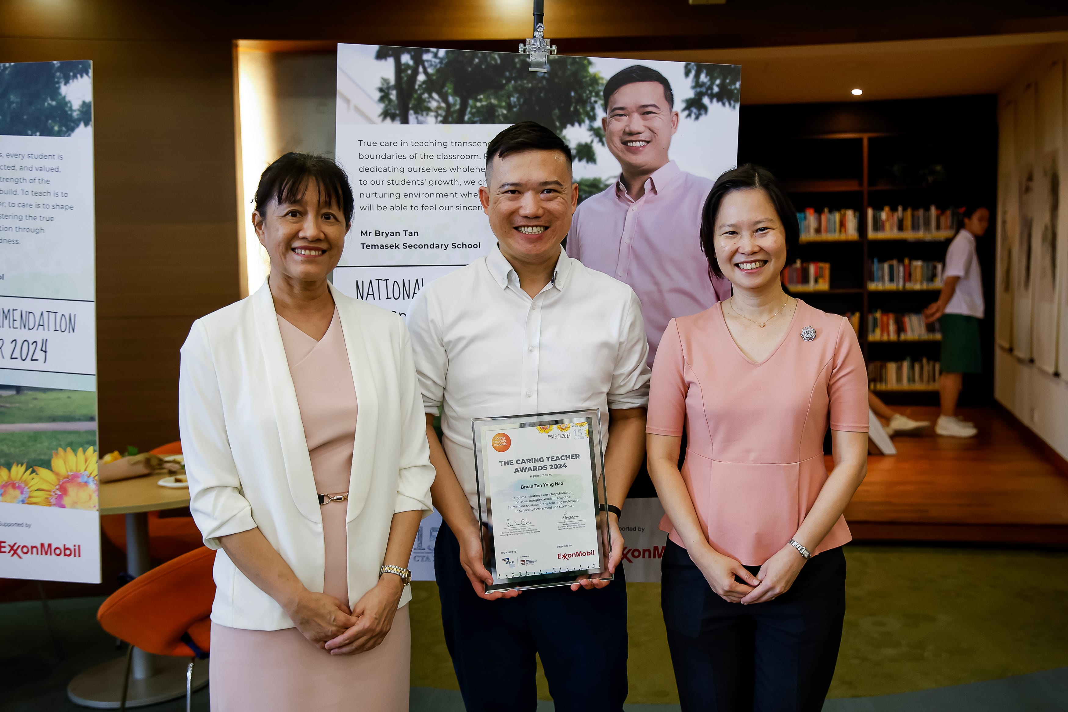 Three adults pose indoors; man in center holds a framed certificate, with award posters and bookshelves behind.