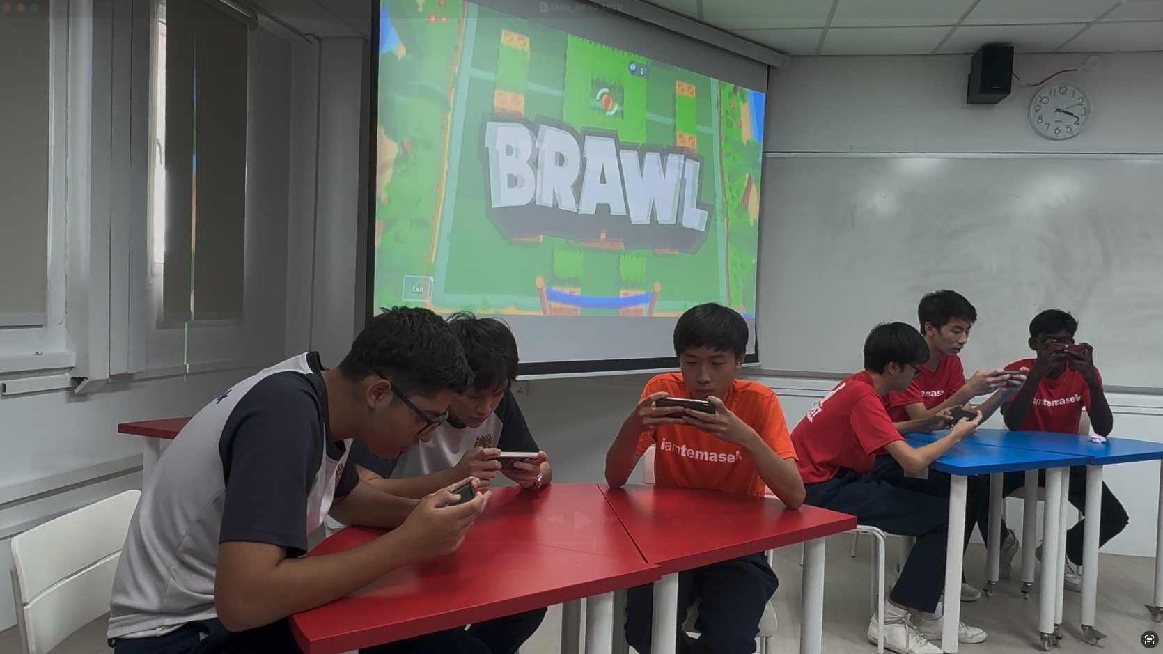 Five students play Brawl Stars on phones in a classroom with the game projected on a screen behind them.