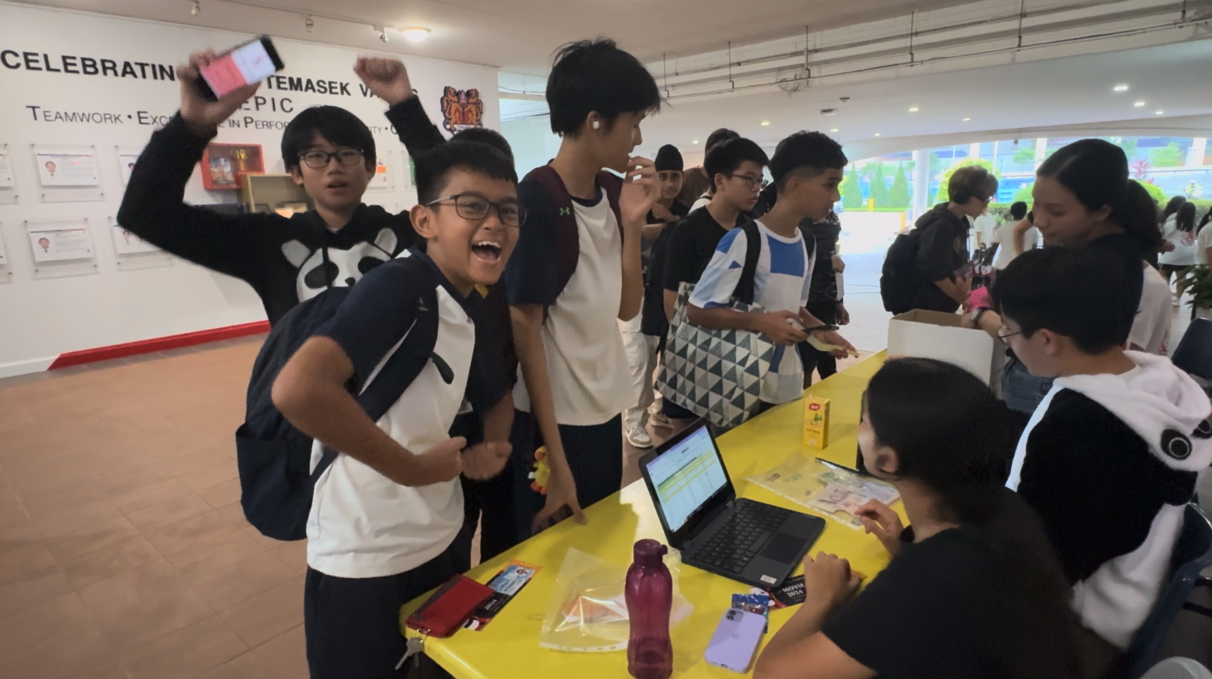 Students gather around a yellow table with a laptop at a school event, with one boy smiling and another raising a phone.