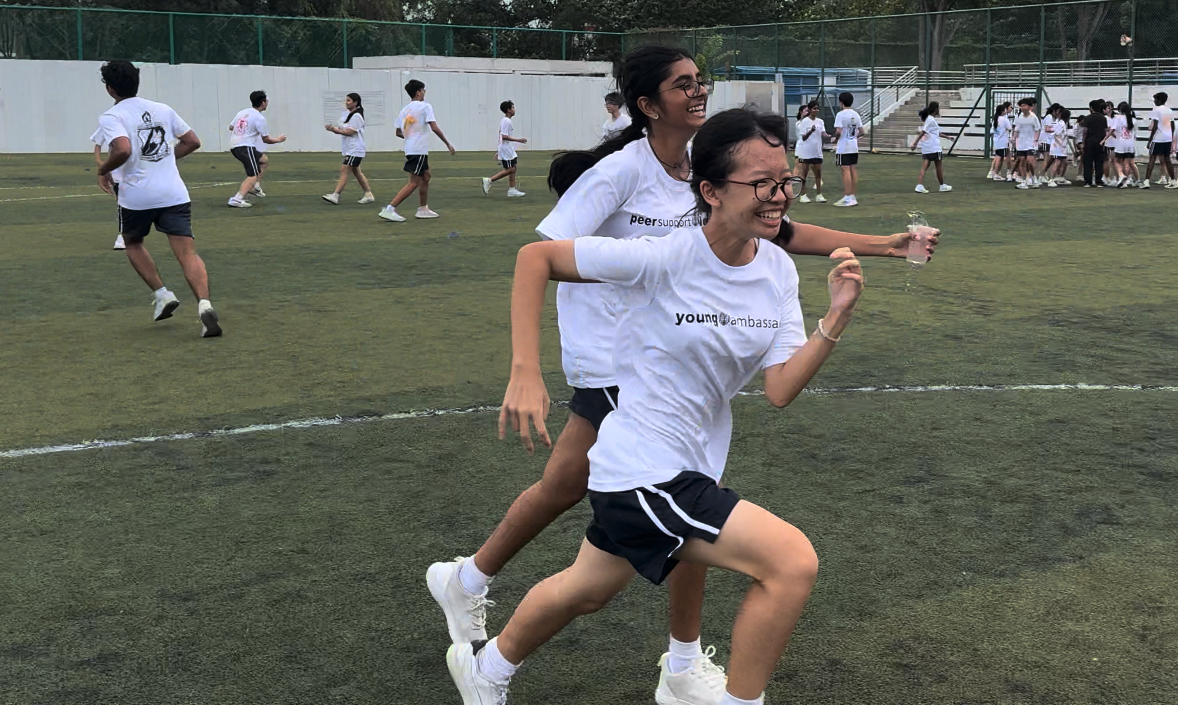 Two smiling girls in white T-shirts run together on a grass field during an outdoor school activity.