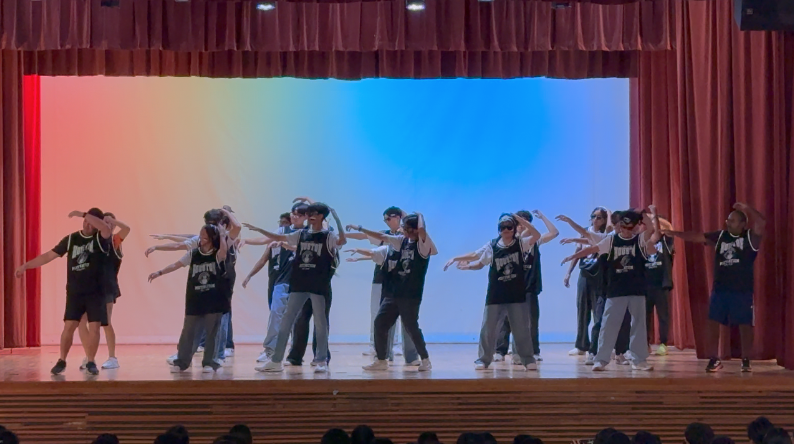 A group of dancers in black jerseys perform on a stage with red curtains and a colorful lit backdrop.
