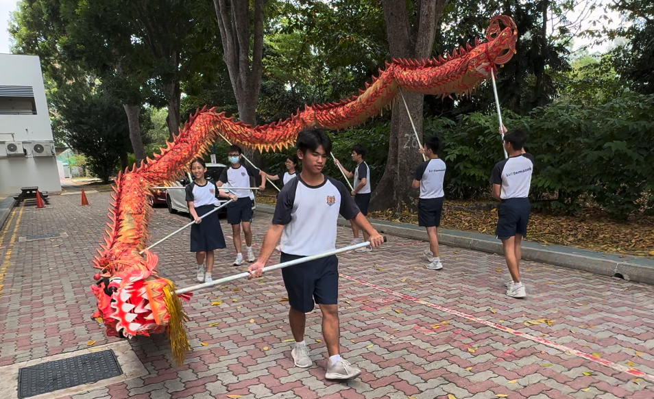 Students in uniforms perform a traditional Chinese dragon dance outdoors, carrying a long red dragon on poles.
