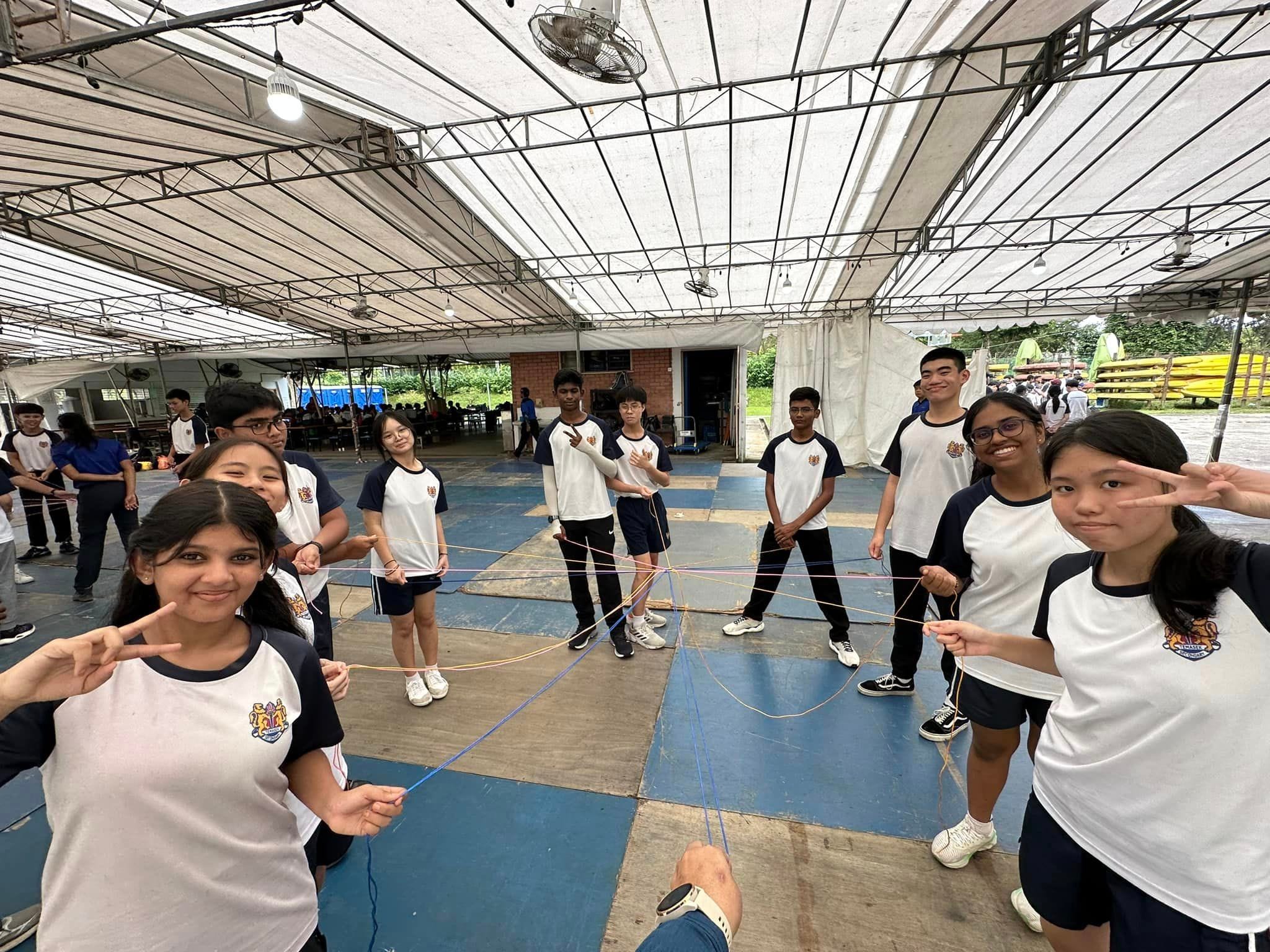 Students in matching uniforms hold colored strings in a team activity under a large open tent.