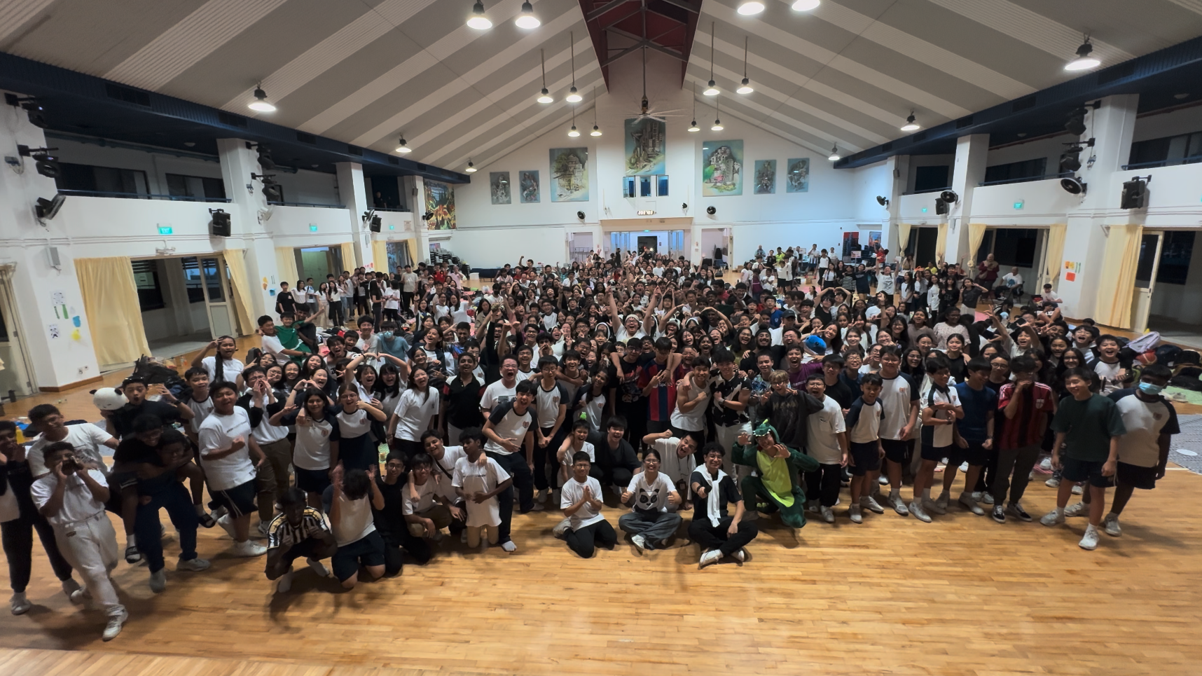 Large group of students posing and cheering together inside a school hall with a wooden floor and high ceiling.