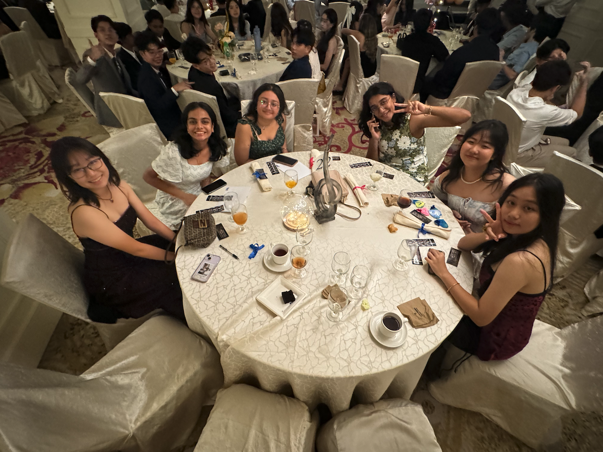 Six young women in formal attire smile at a decorated round banquet table in a crowded ballroom.