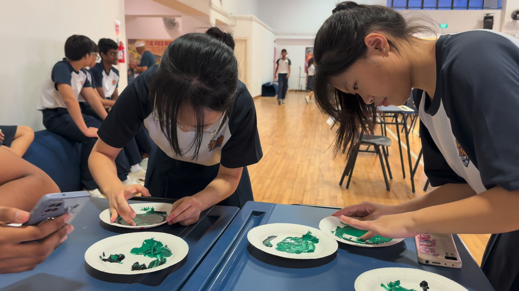 Two school students in uniforms lean over a table, pressing green paint onto white paper plates indoors.