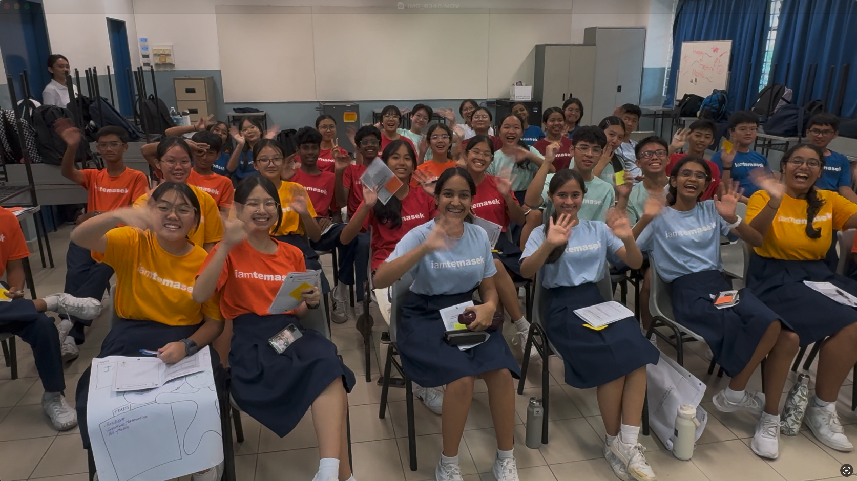A large group of students wearing colorful iamtemasek shirts wave and smile in a classroom.