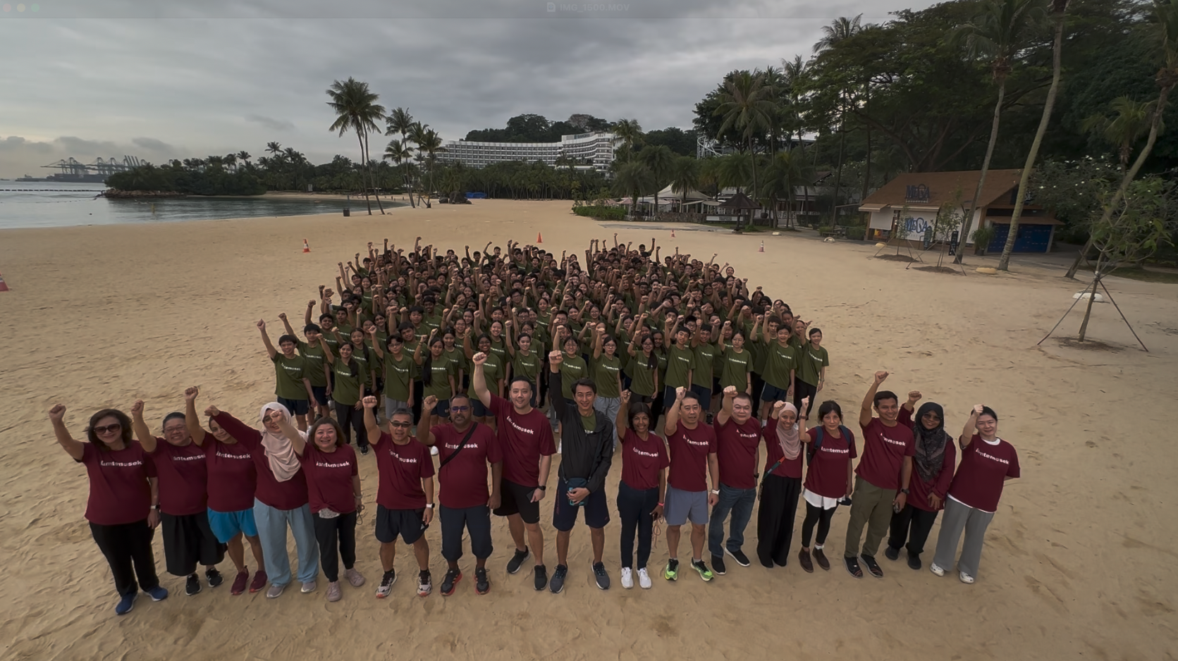 Large group in matching red and green shirts raising fists on a sandy beach with palm trees and a hotel behind them.