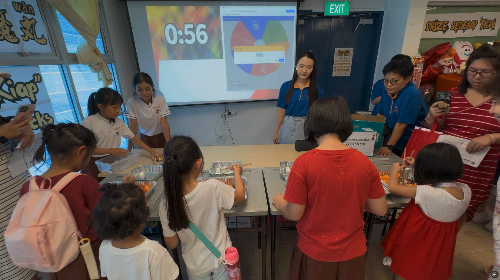 Children and adults gather around tables doing a timed activity, with a countdown of 0:56 displayed on a projector screen.