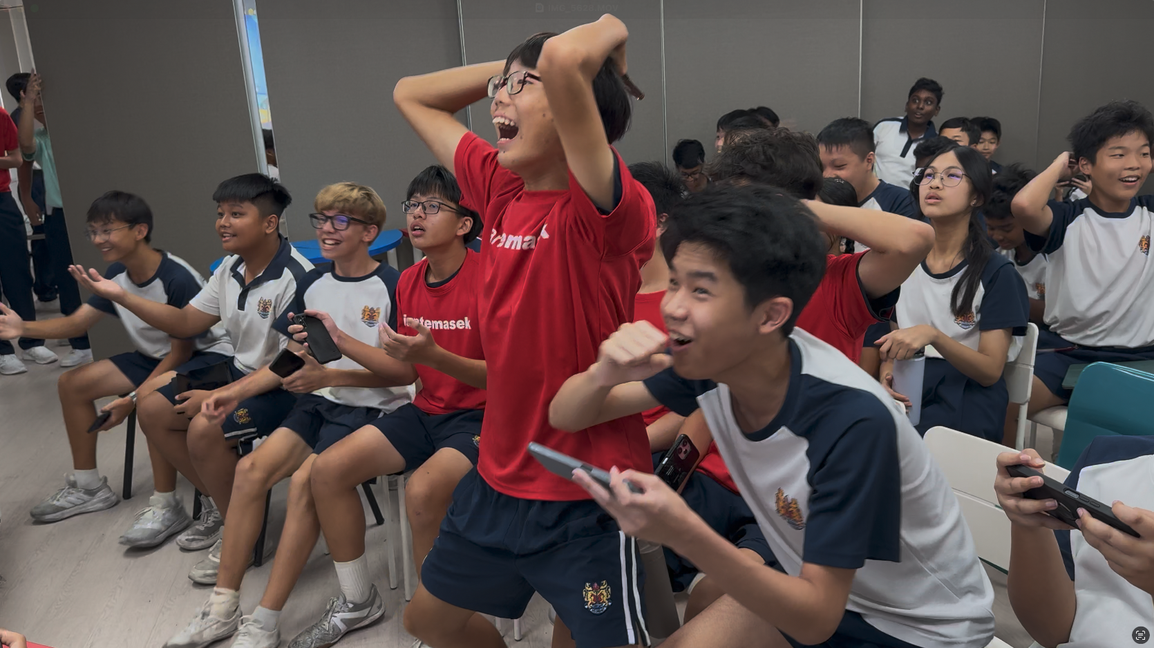 A group of excited school students in uniforms react with shock and joy, some holding phones and raising hands.
