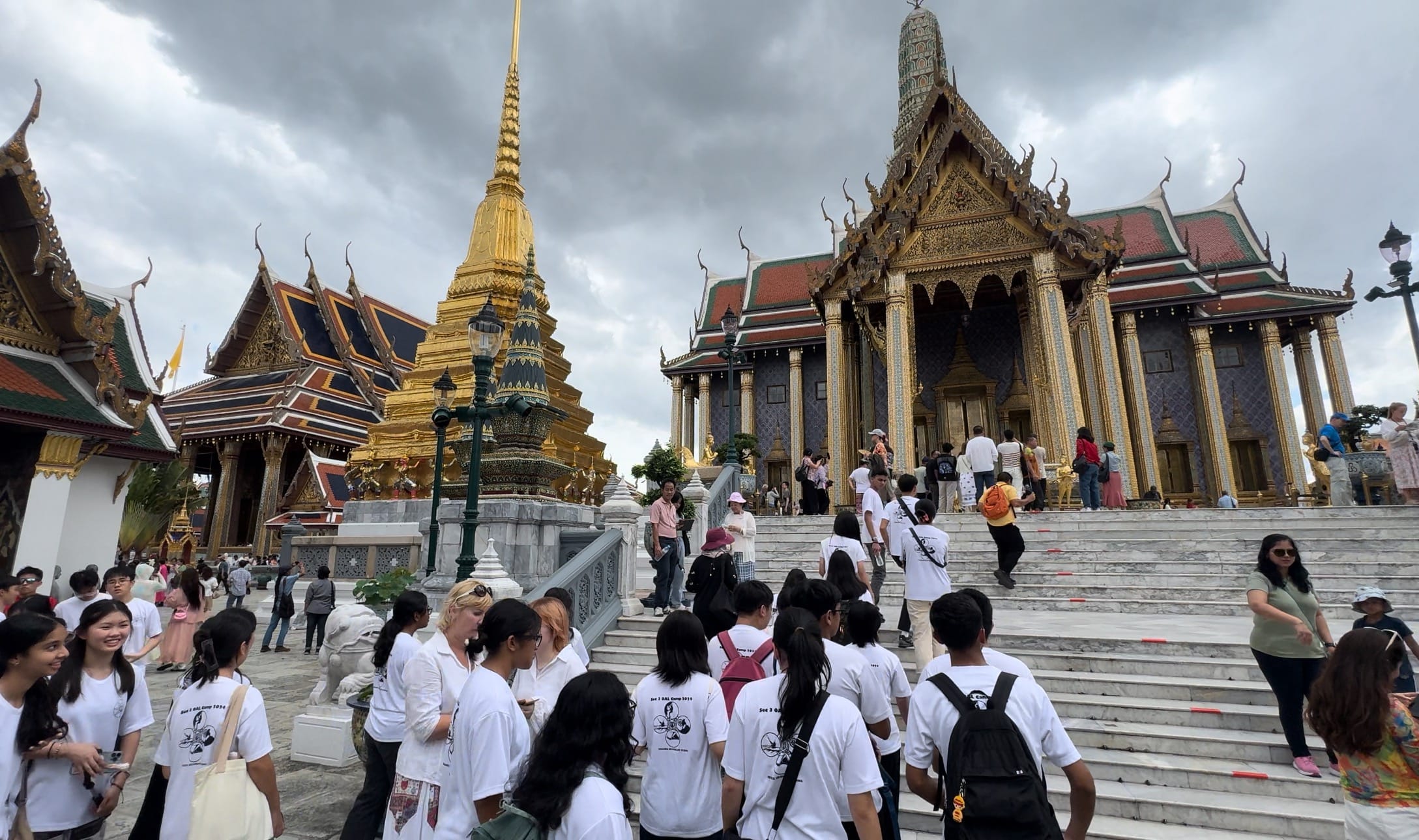 A crowd of visitors in white shirts walks up wide steps toward an ornate Thai temple with a golden spire.