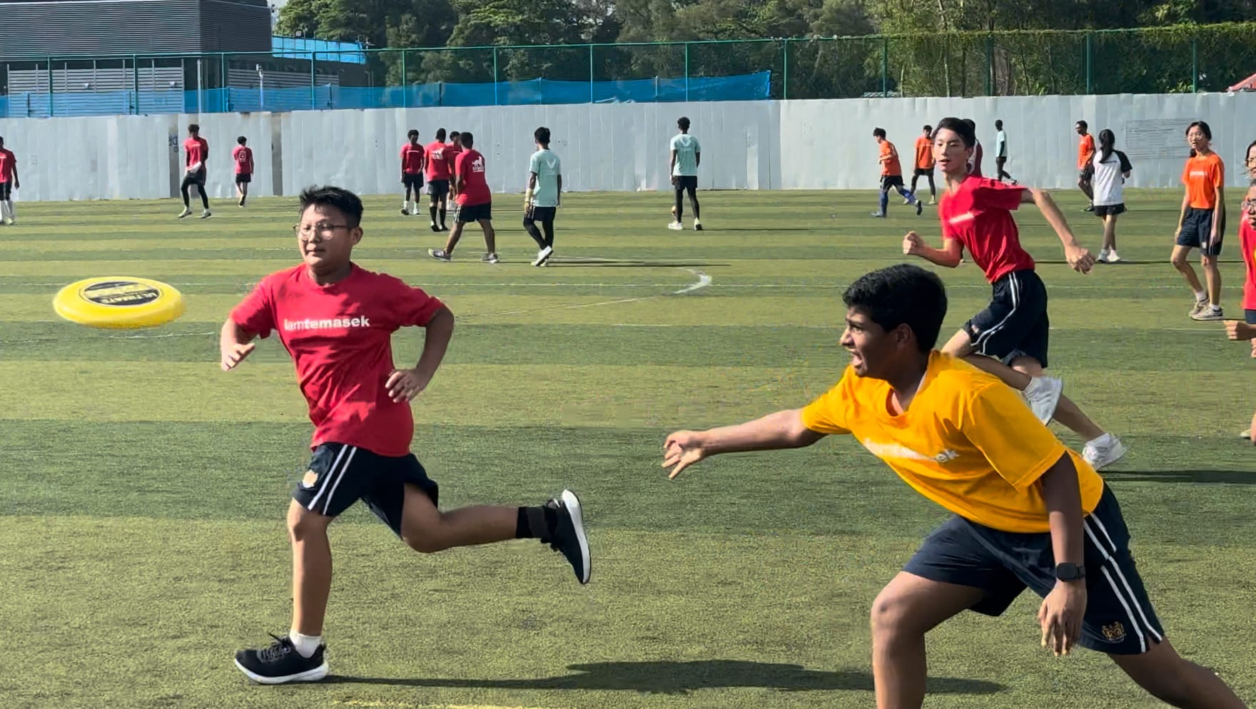 Students play ultimate frisbee on a green field, with a yellow disc visible mid-air near players in red and yellow shirts.