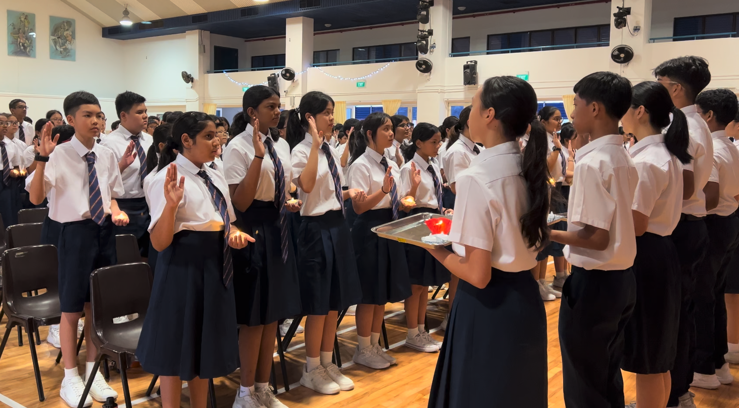 Students in white shirts and navy uniforms raise their hands in a pledge ceremony as a girl holds a candle tray.