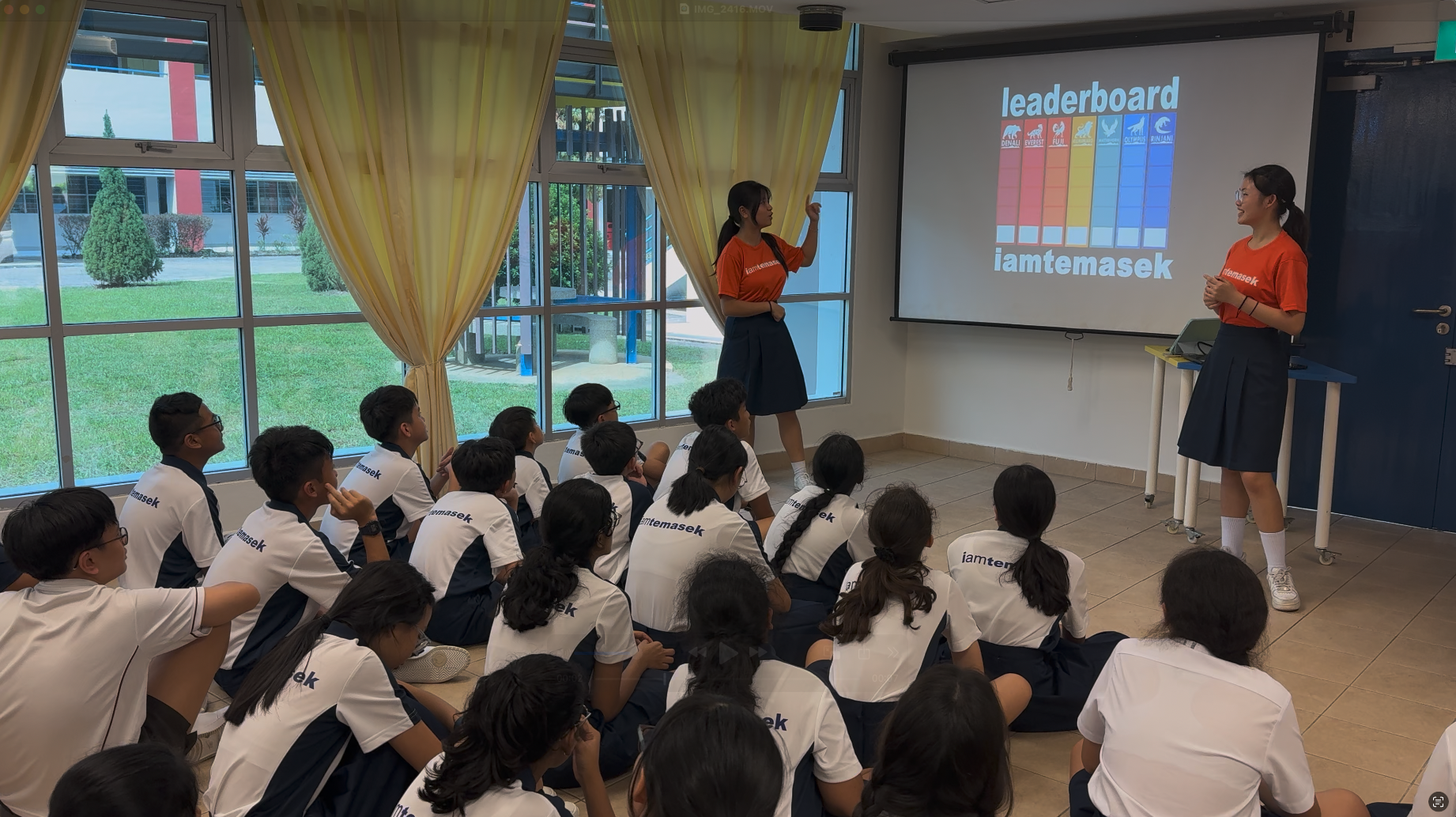 Two students in orange shirts present a leaderboard slide to uniformed pupils sitting on the floor in a classroom.