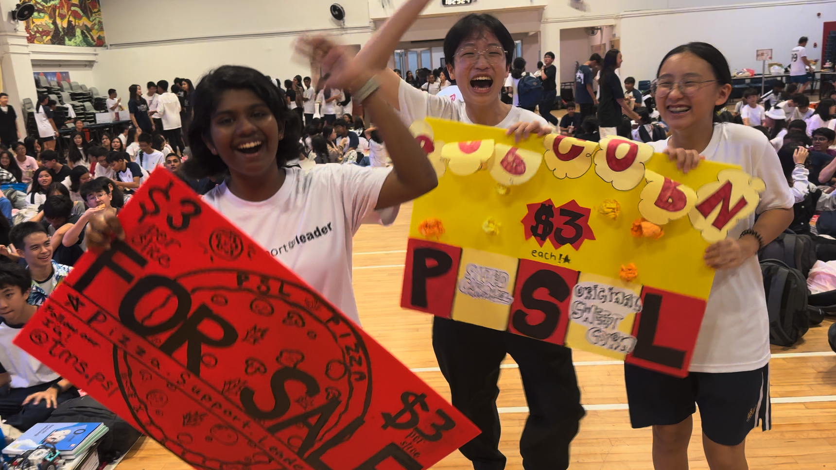 Three students cheer in a gym holding handmade signs advertising popcorn and pizza for $3 each.
