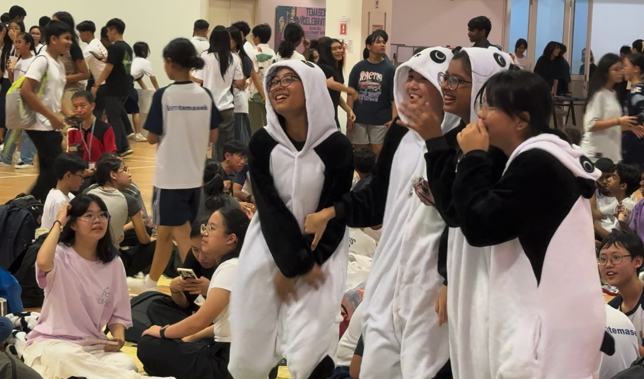 Three smiling students wear panda onesies at a crowded school event, surrounded by seated and standing peers.