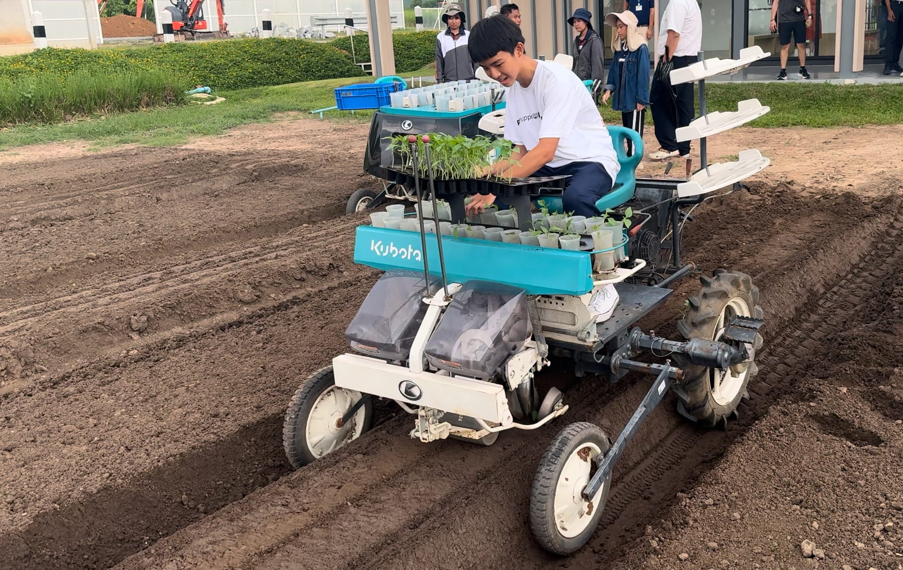 A young man rides a Kubota seedling transplanter across tilled soil rows, with onlookers in the background.