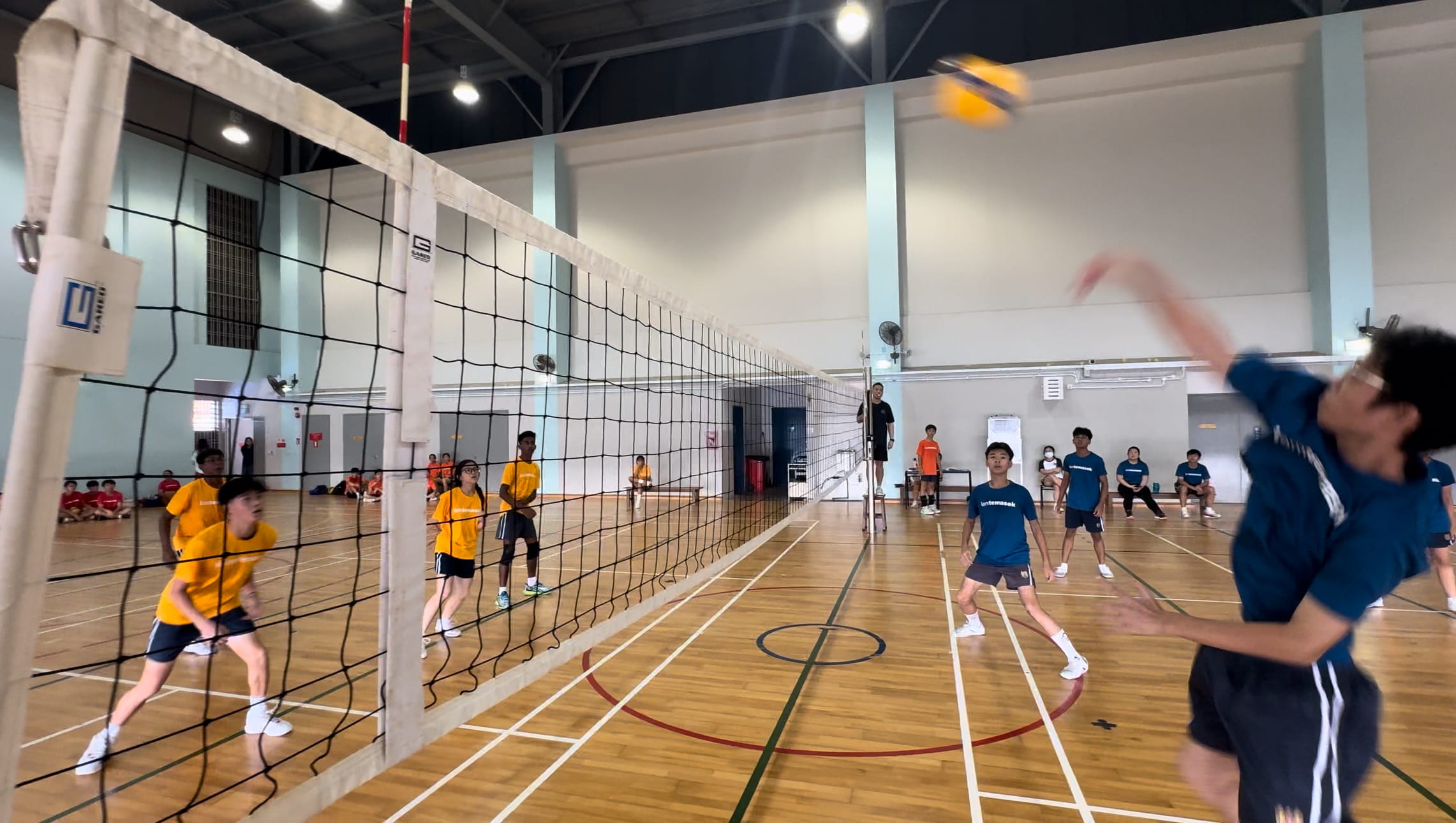 Players in yellow and blue shirts compete in an indoor volleyball match, with a ball mid-air above the net.