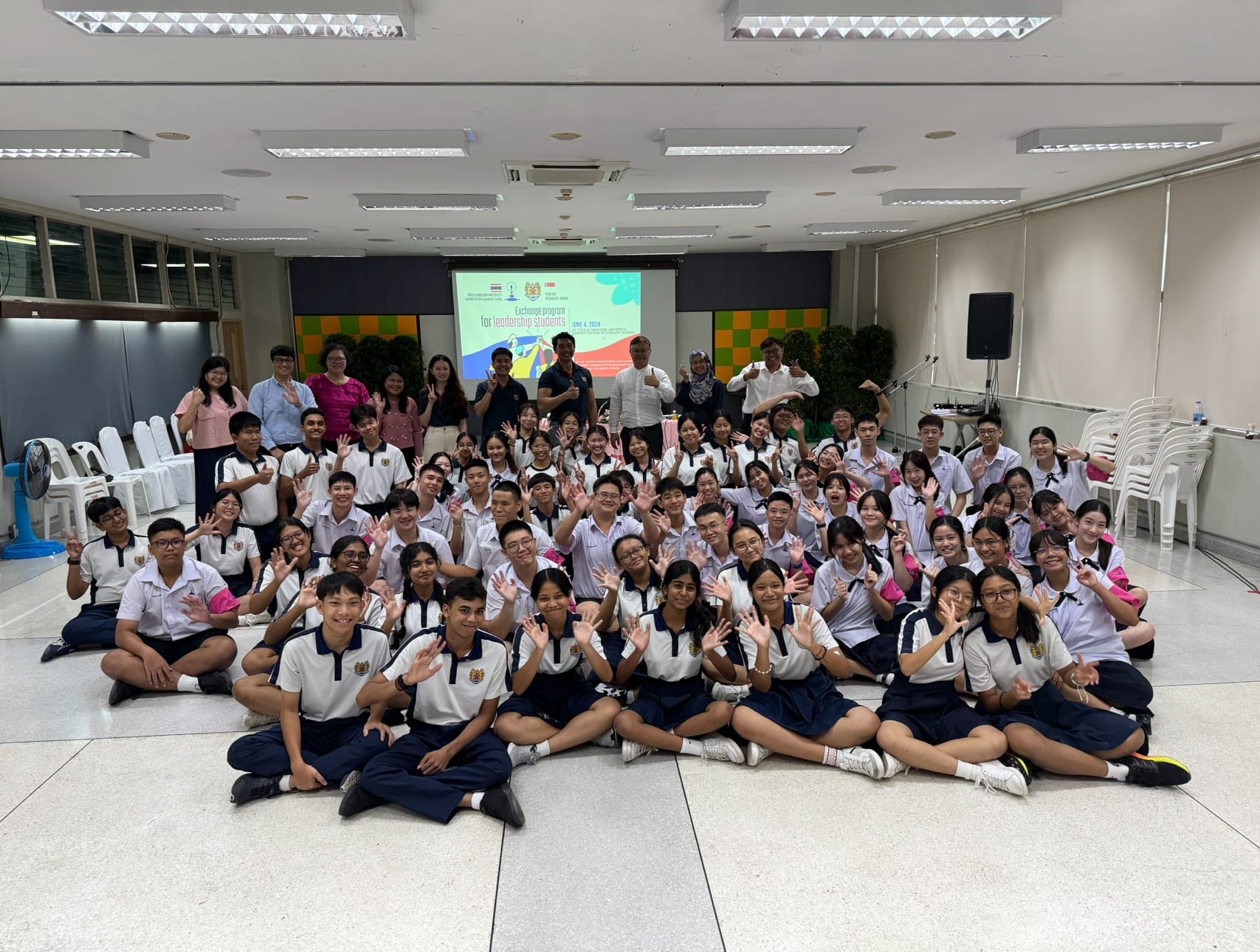 Large group of school students in uniforms posing and waving at a leadership exchange program event indoors.