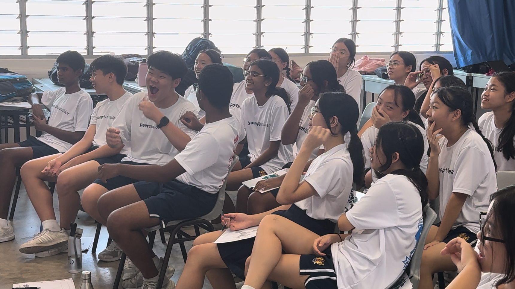 A group of teenagers in white t-shirts sit and laugh together in a bright classroom setting.