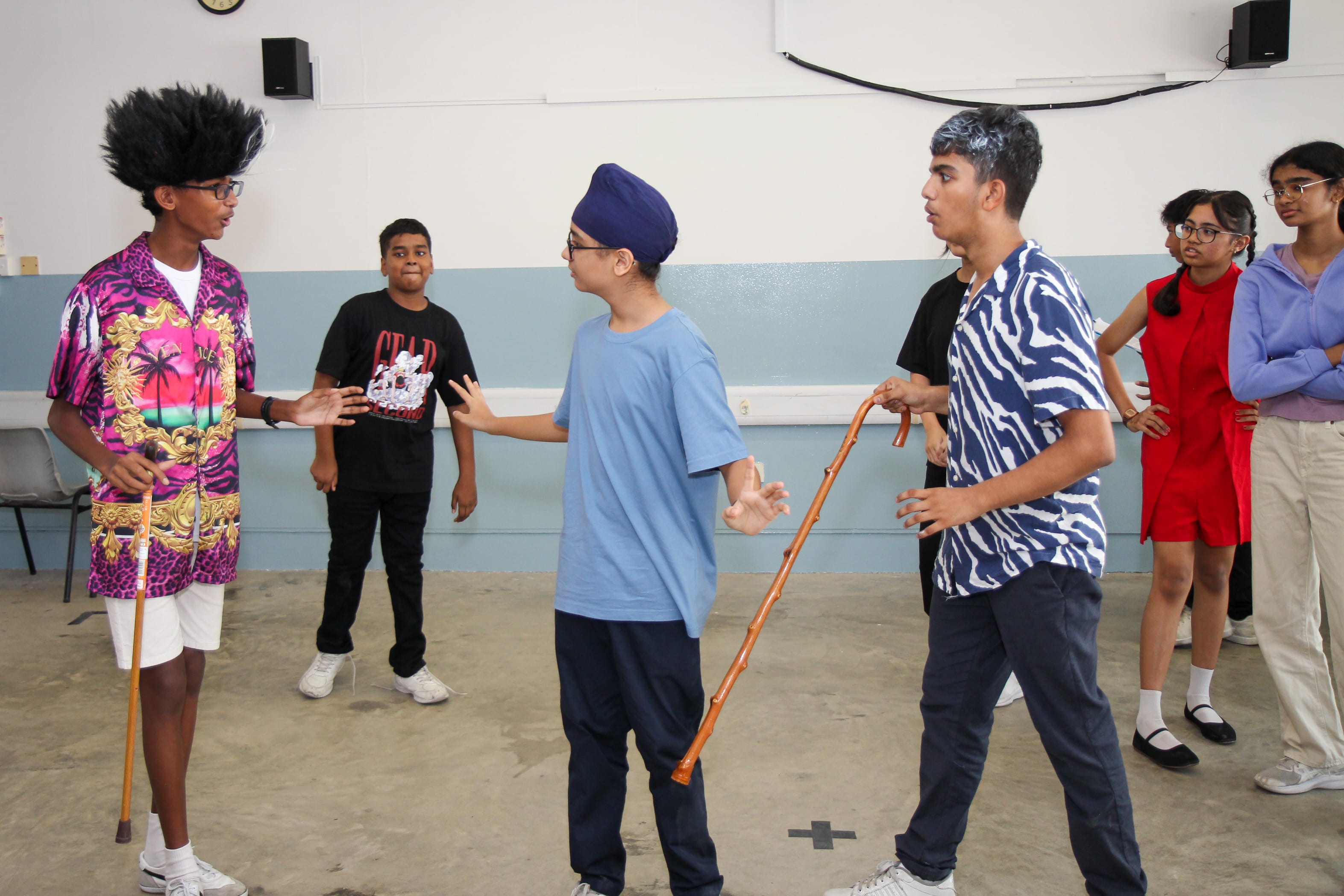 Teens in a room act out a scene; one holds a cane, another gestures, others watch in the background.