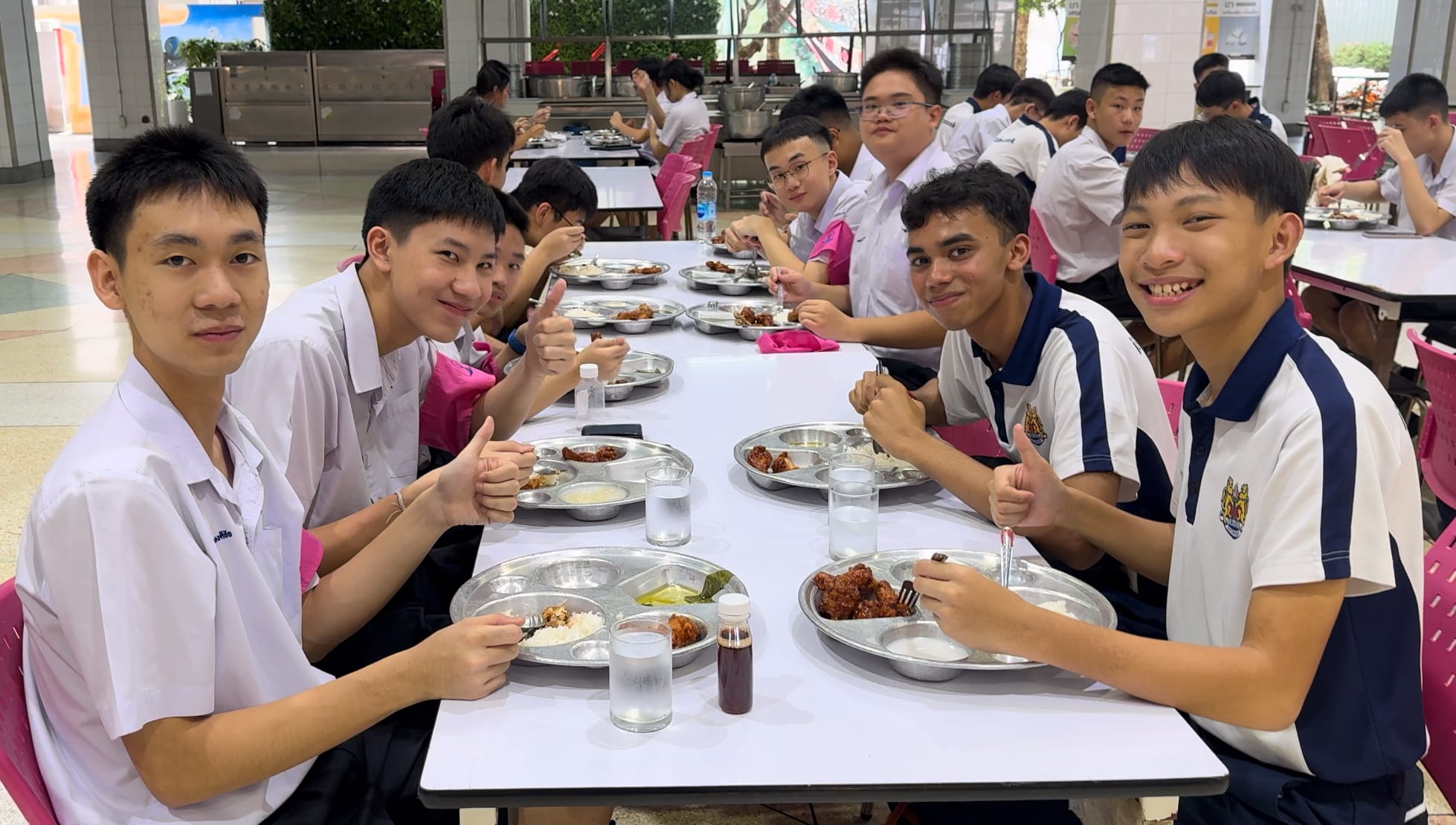 Several teenage students in school uniforms sit at a long table eating meals on metal trays, giving thumbs up.