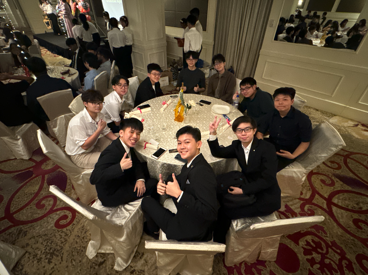 A group of young men in formal wear sit around a round banquet table, smiling and posing at an indoor event.