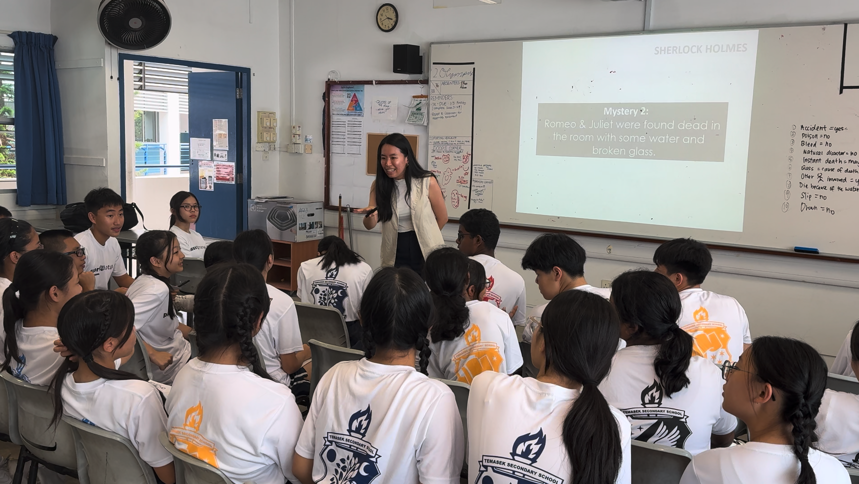 A teacher stands at the front of a classroom presenting a Sherlock Holmes mystery slide to uniformed secondary school students.