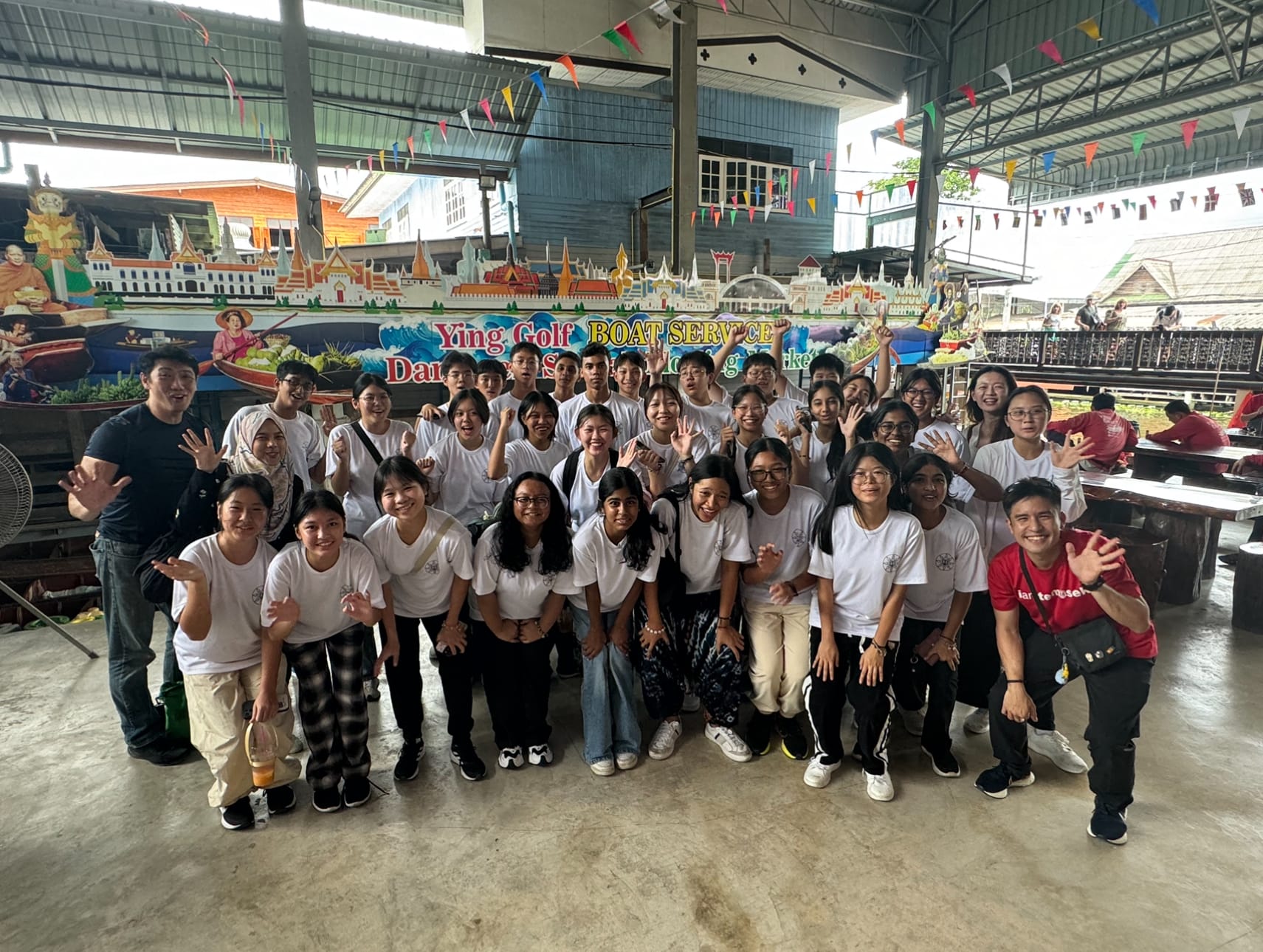 A large group of students in white shirts poses and waves at a Thai floating market boat service pavilion.