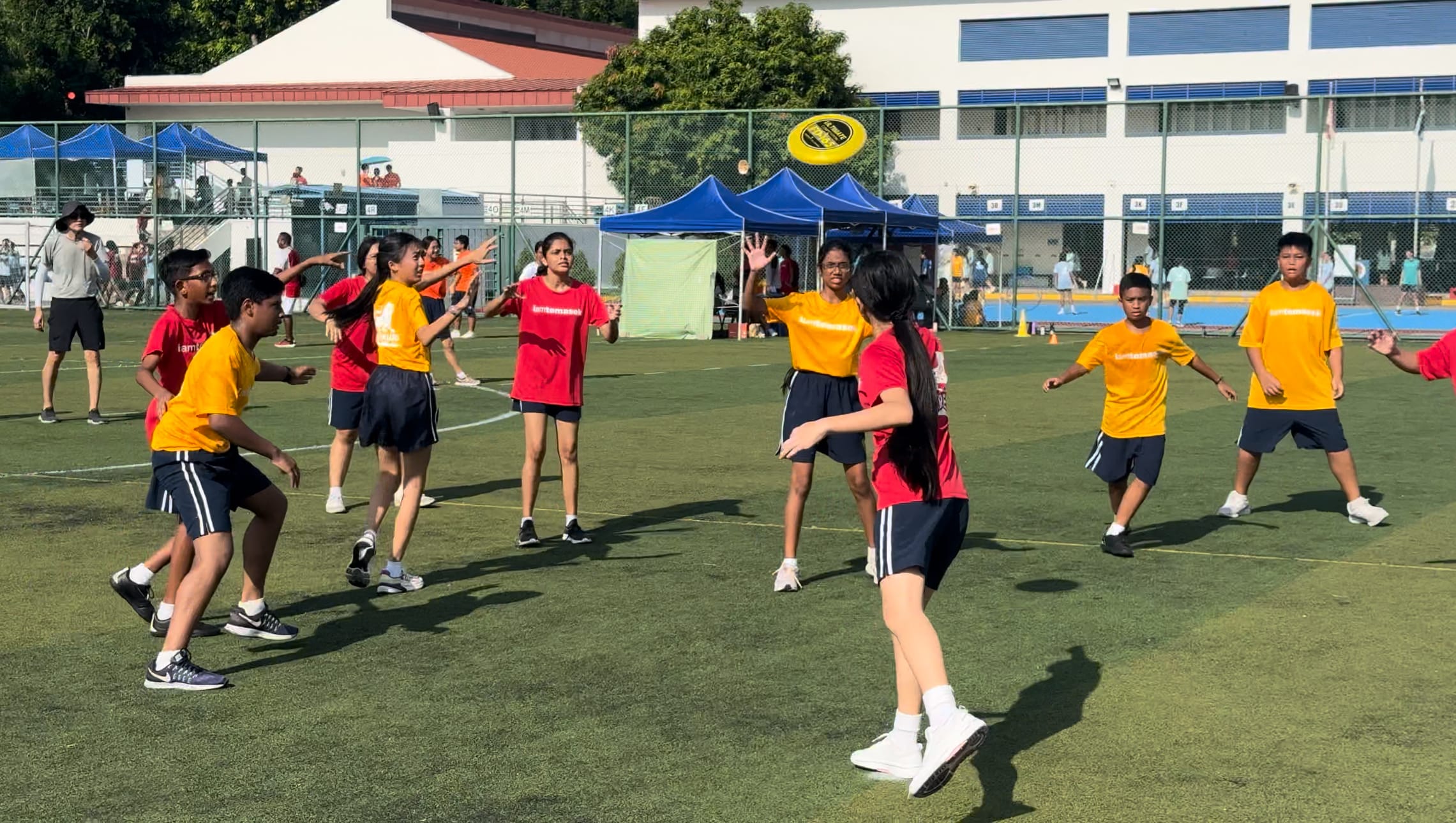 Students in red and yellow shirts play ultimate frisbee on a green field with blue canopy tents in the background.