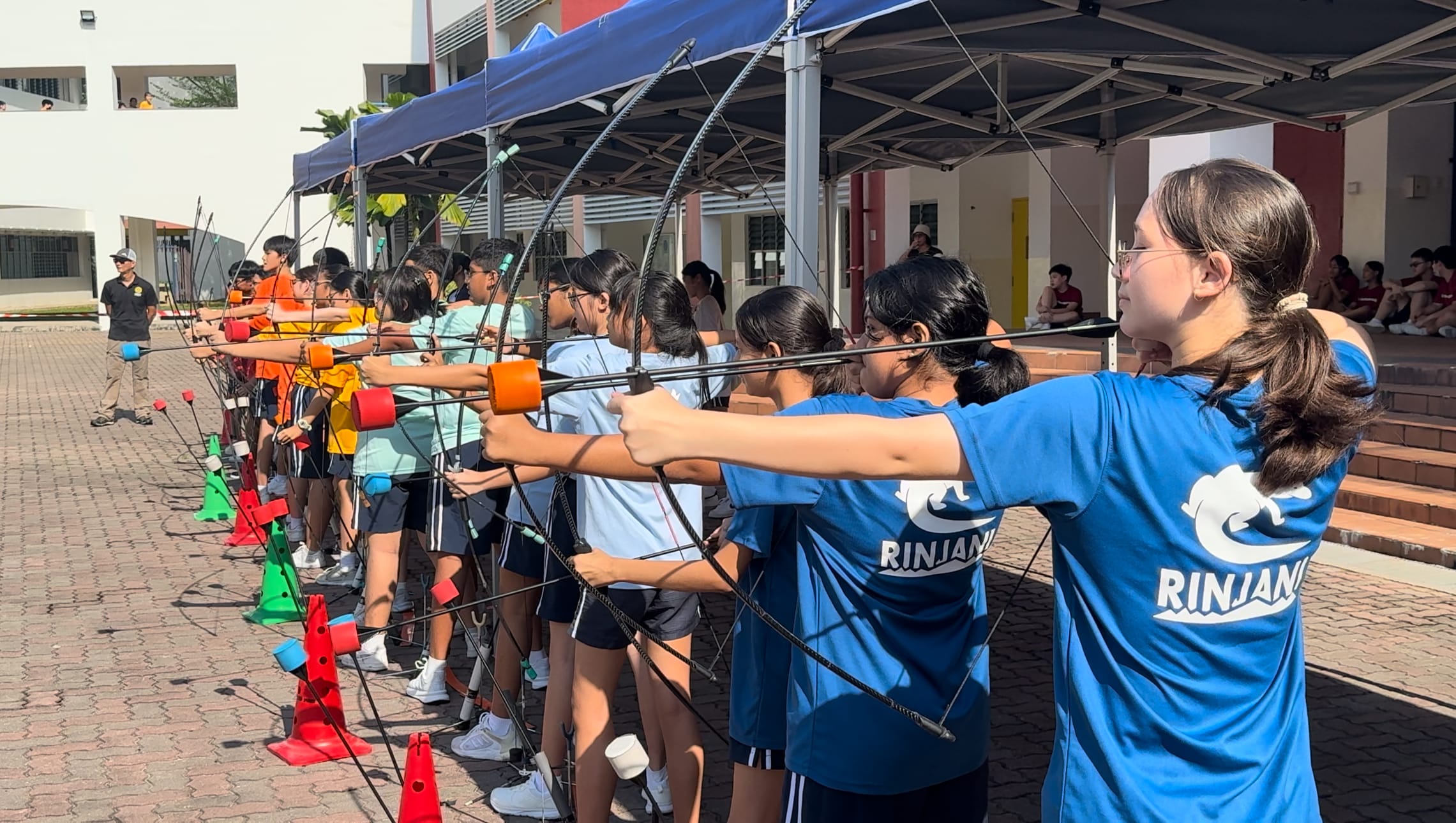 A row of students in colorful shirts aim bows during an outdoor archery session under a blue canopy.