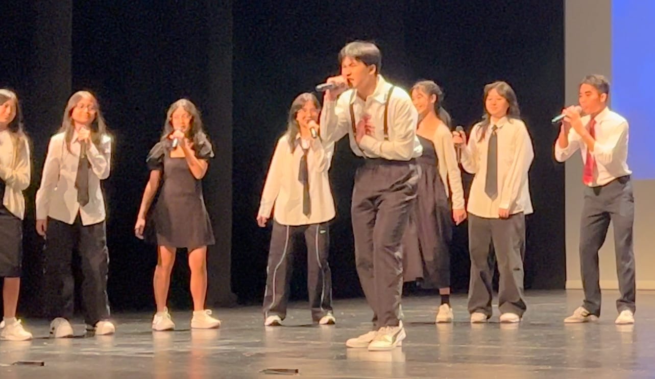 A group of young performers singing into microphones on a dark stage, wearing white shirts and ties.