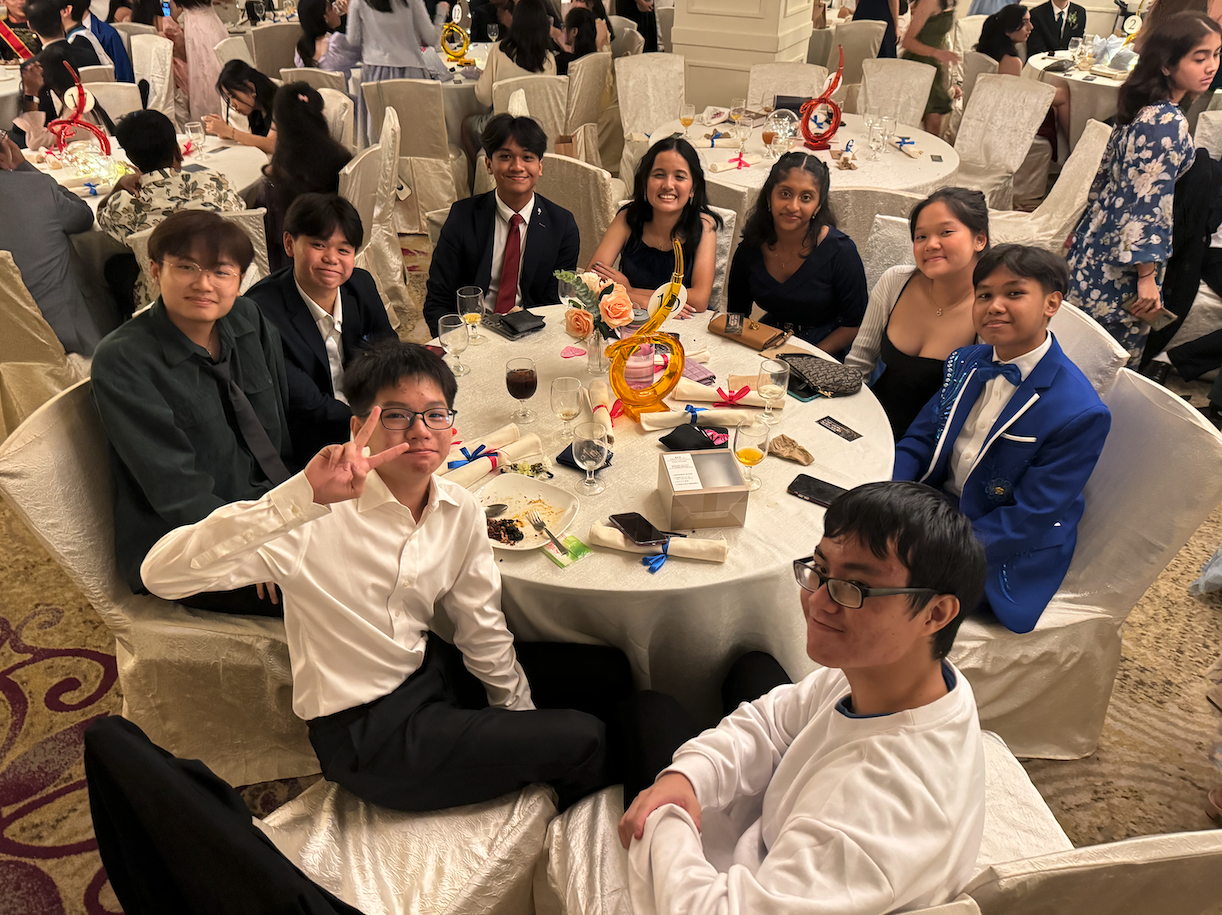 A group of formally dressed teenagers sitting around a round banquet table, smiling at the camera.