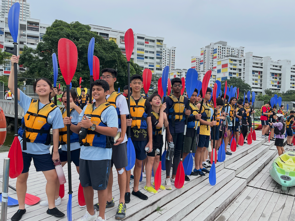 A group of young students in orange life jackets hold red and blue kayak paddles on a waterfront dock.