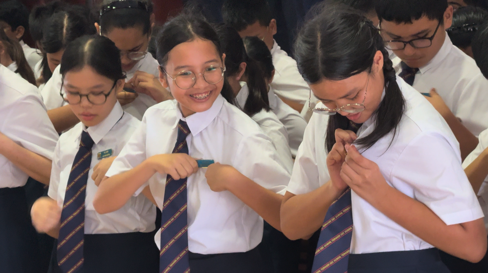 Students in white shirts and striped ties adjusting their ties, with one girl smiling broadly at the camera.