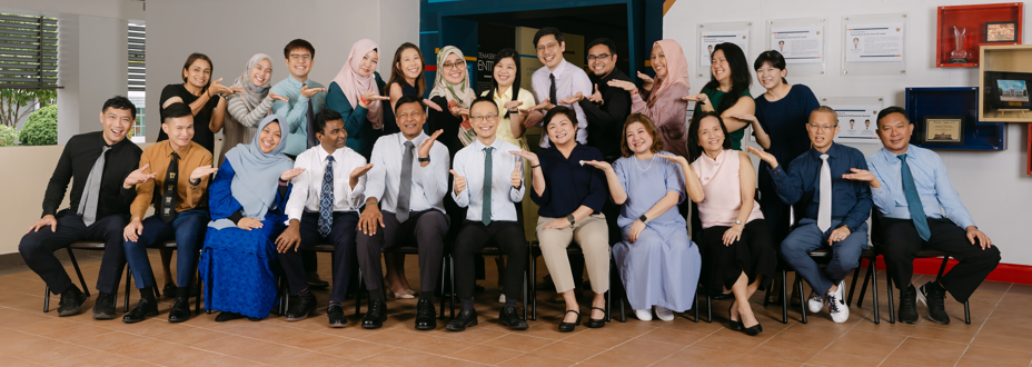 A large group of about 25 people posing for a photo in front of an "Our Temasek Story" display.