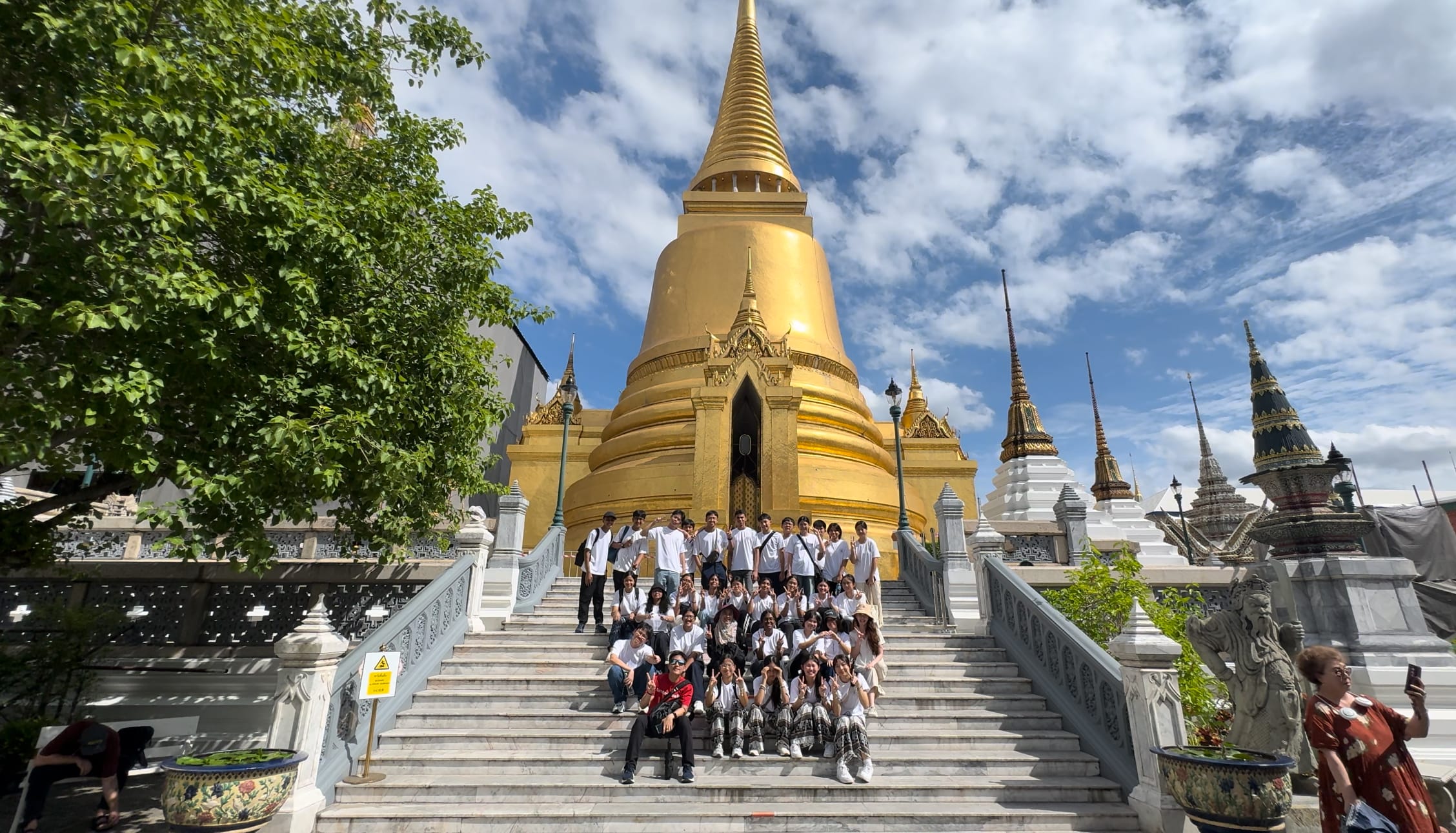 A large group poses on white marble steps before a towering golden Buddhist chedi under a blue sky.
