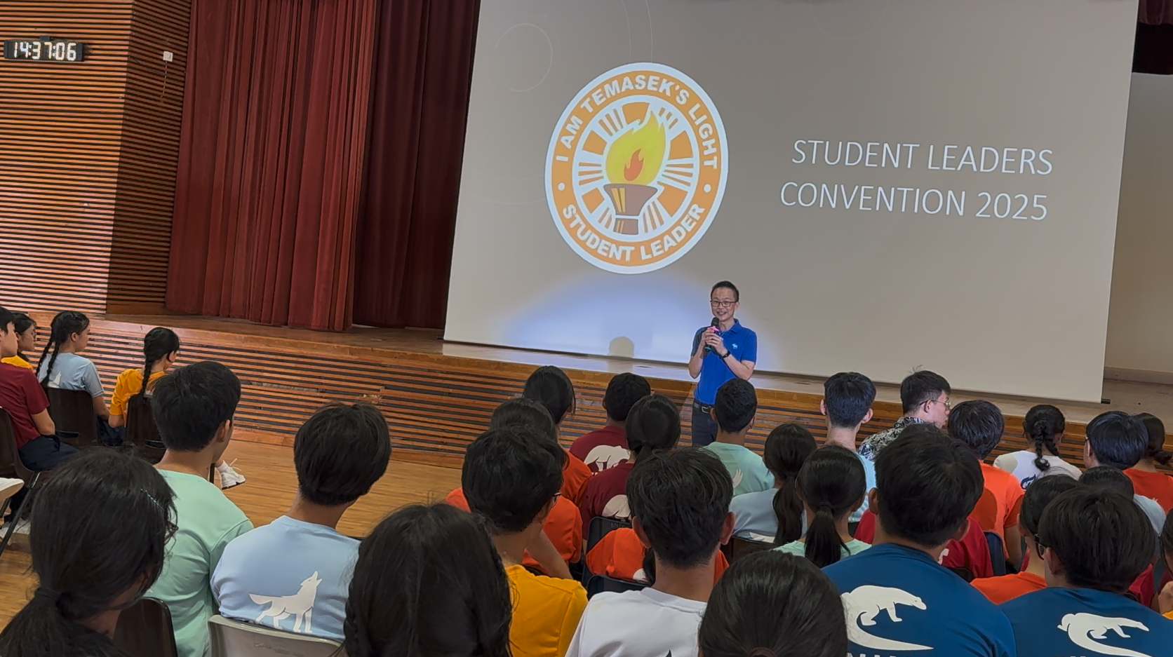 A man in a blue shirt speaks at the Temasek Student Leaders Convention 2025 before a seated audience in a hall.