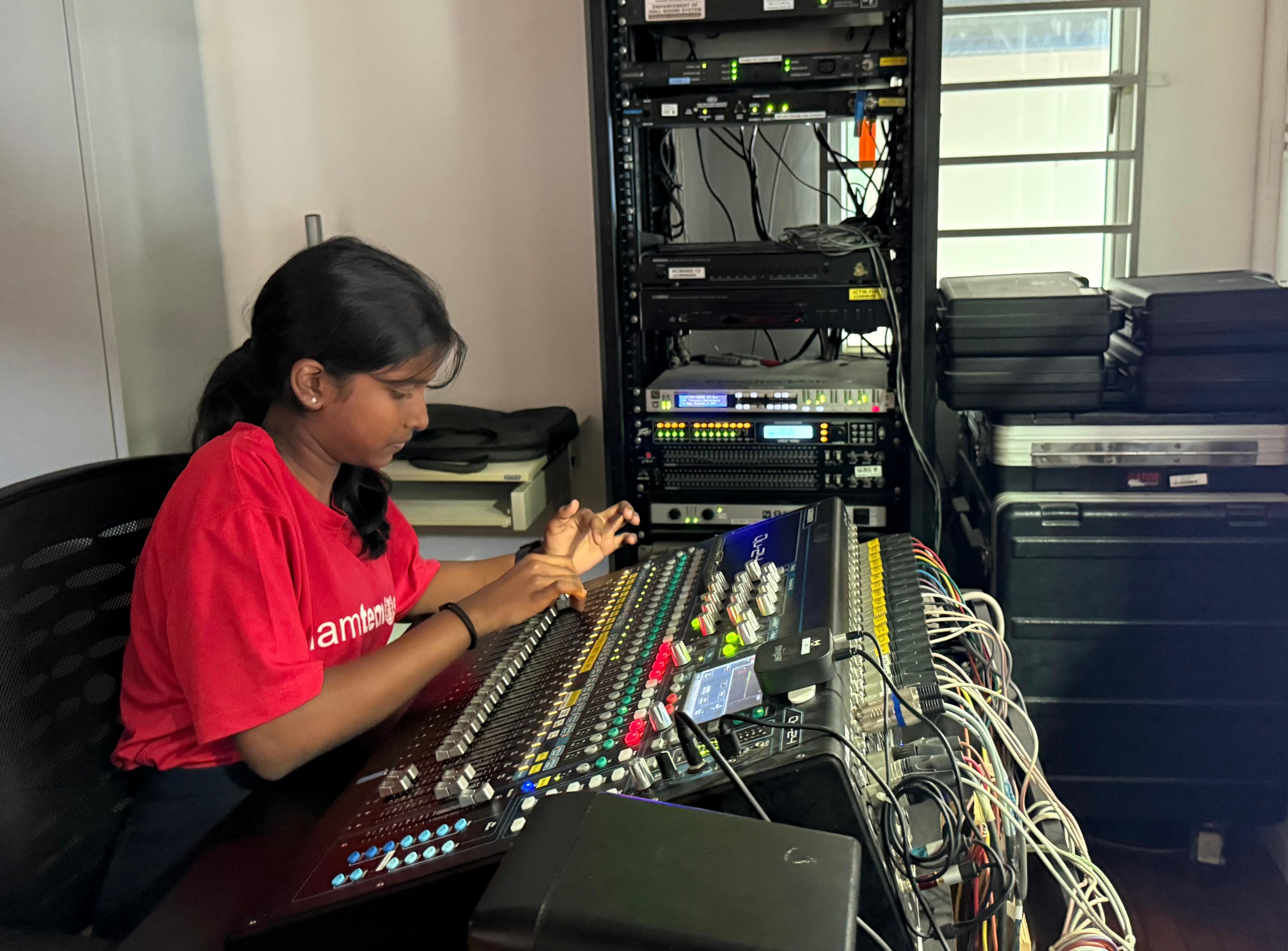A young woman in a red shirt operates a large audio mixing console in a room with server rack equipment.