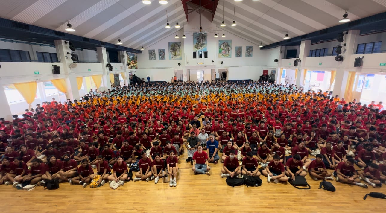 Large group of students in red, orange, and teal shirts seated on a school hall floor for a group photo.