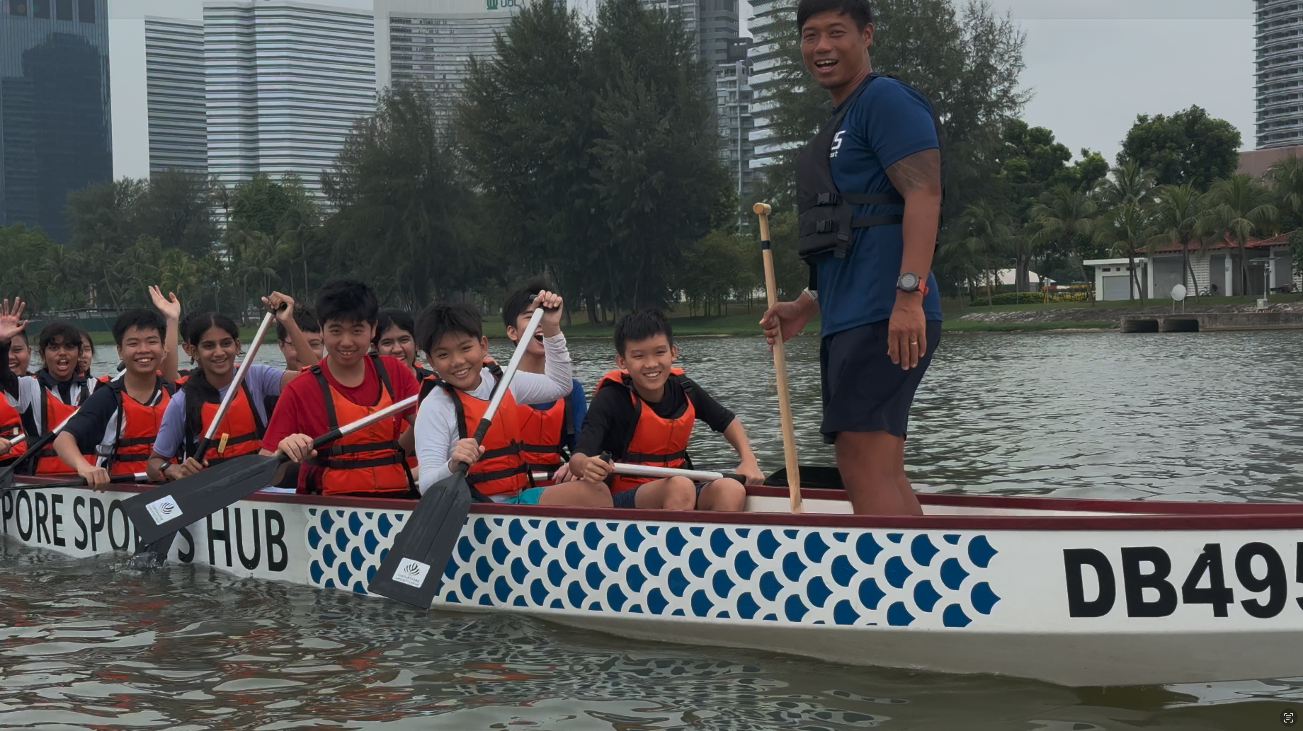 A group of smiling children in orange life vests paddle a Singapore Sports Hub dragon boat guided by a standing adult.