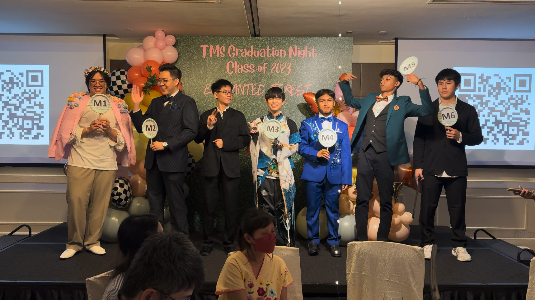 Six formally dressed students hold numbered paddles on stage at TMS Graduation Night Class of 2023 Enchanted Forest event.